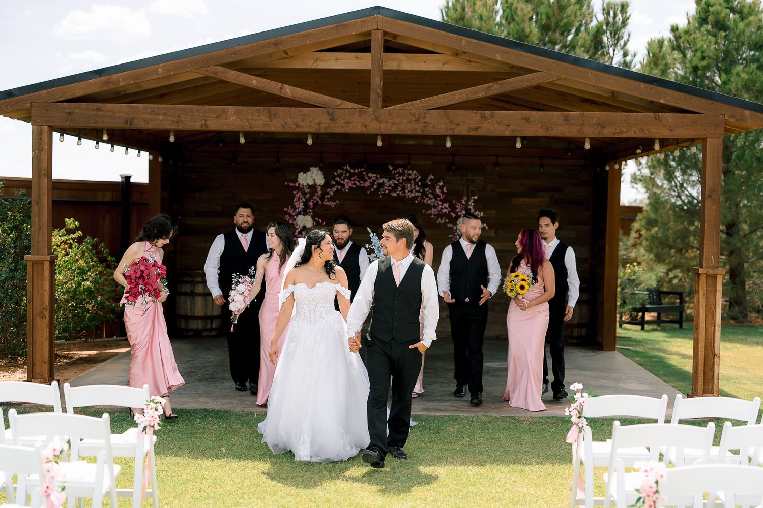 Wedding party exits rustic wooden pavilion, bridesmaids in pink dresses and groomsmen in black vests, bride and groom lead procession.