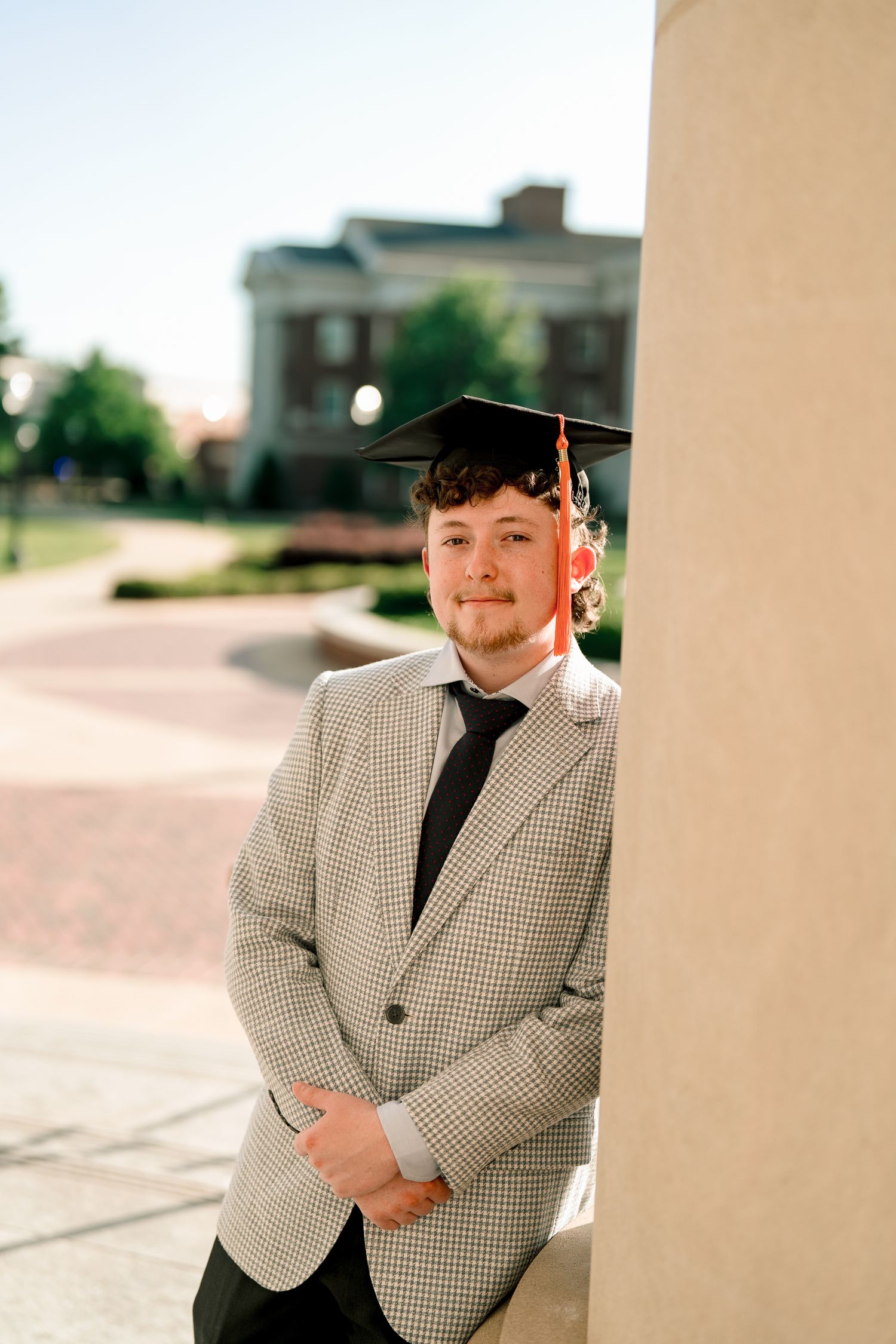 Smiling graduate in mortarboard cap and grey blazer leans against white column on UA campus.