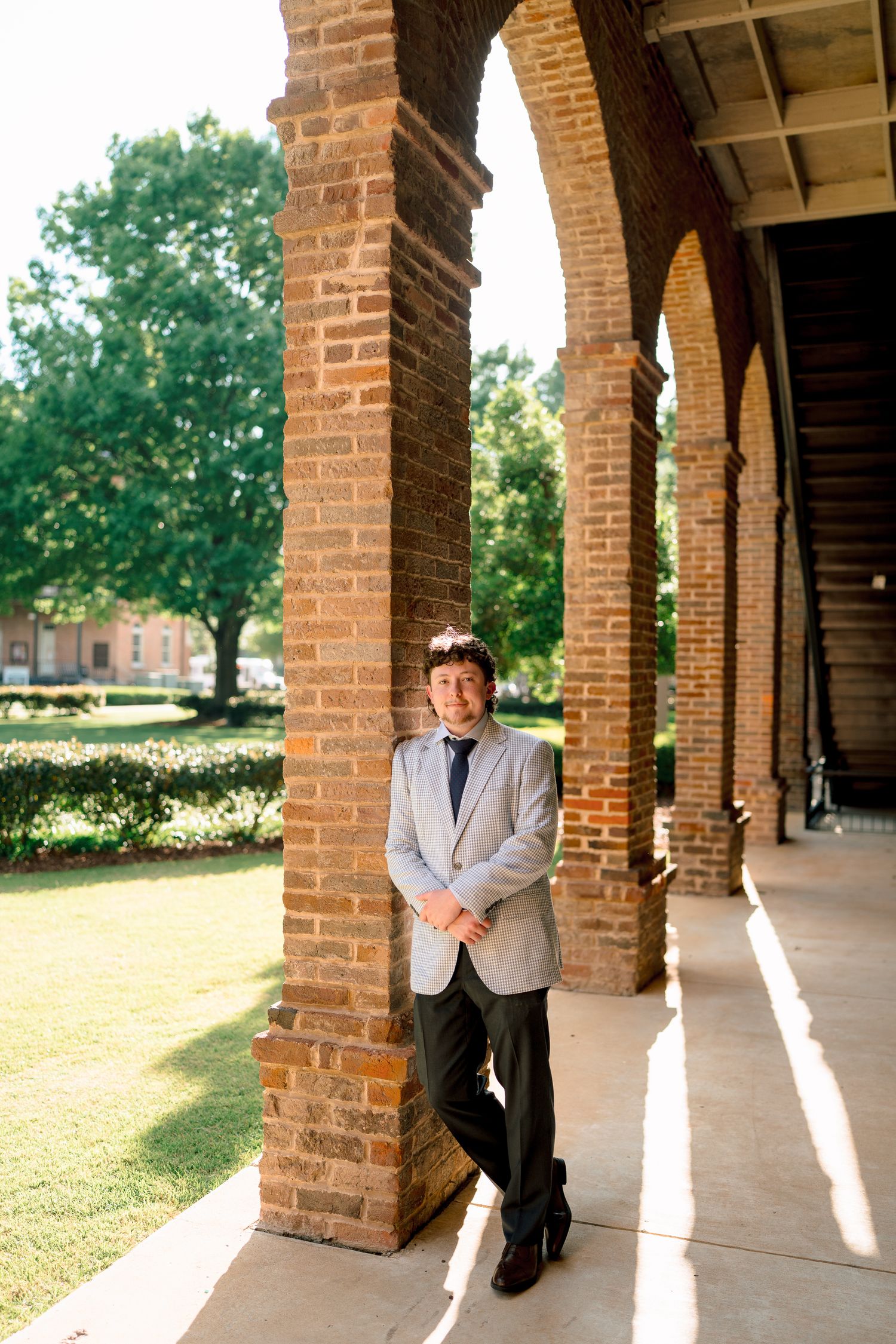 Smiling young man in gray blazer and tie leans against brick arch column in sunlit outdoor corridor.