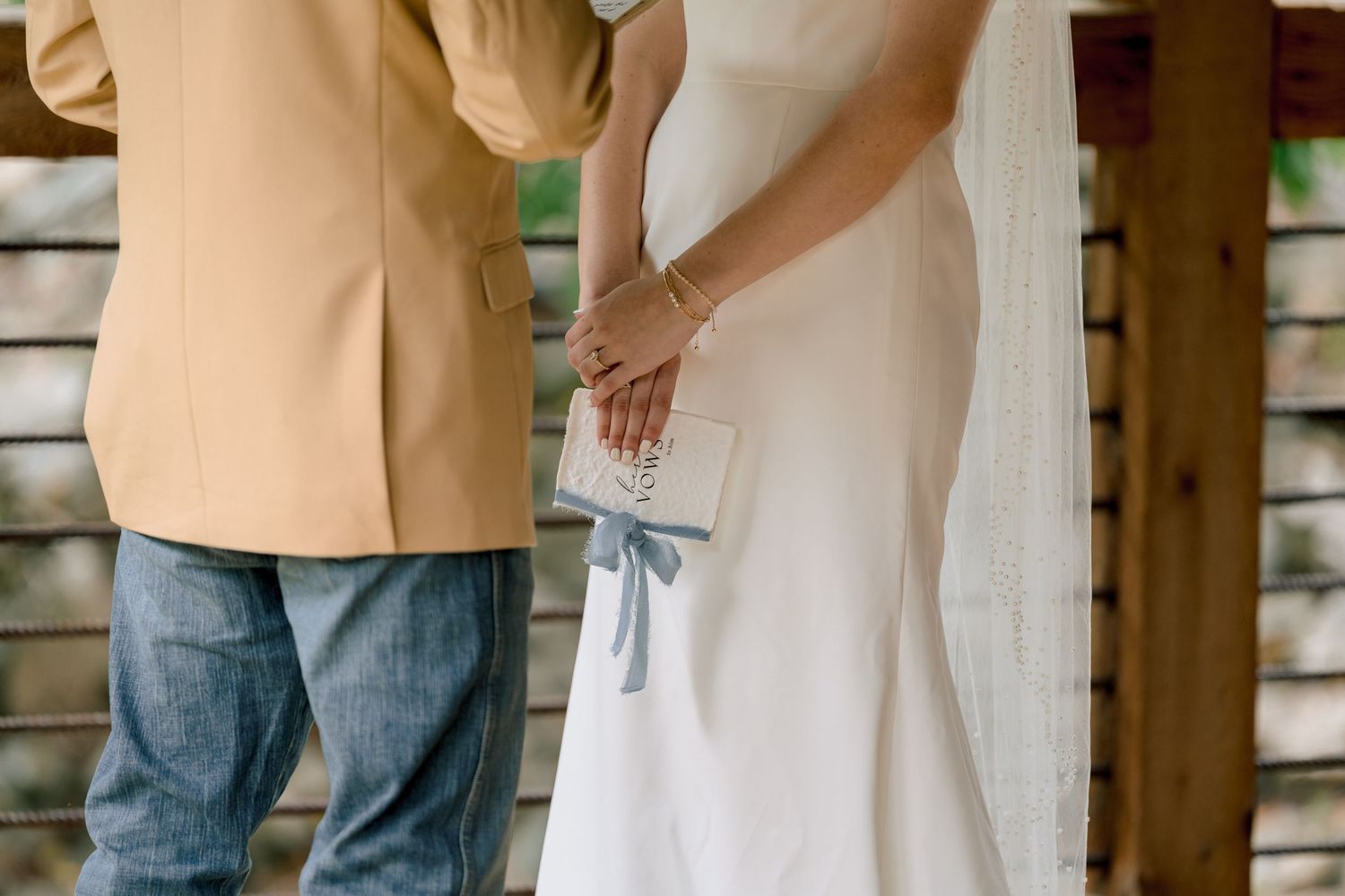 Bride in white gown holding blue ribbon gift box stands beside groom in tan blazer and jeans at outdoor wedding.