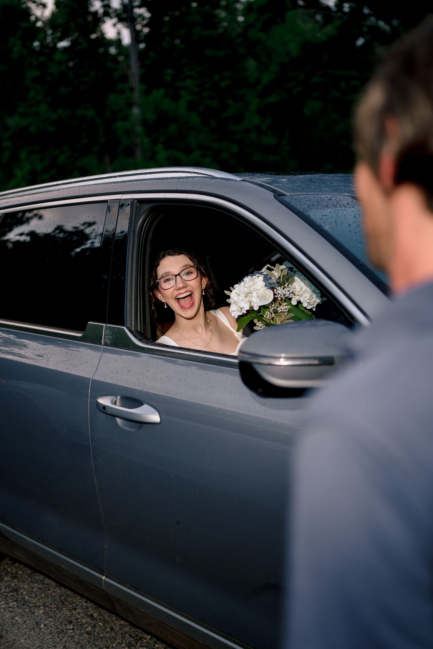 Joyful bride leaning out car window holding white bouquet smiling at groom during wedding getaway.