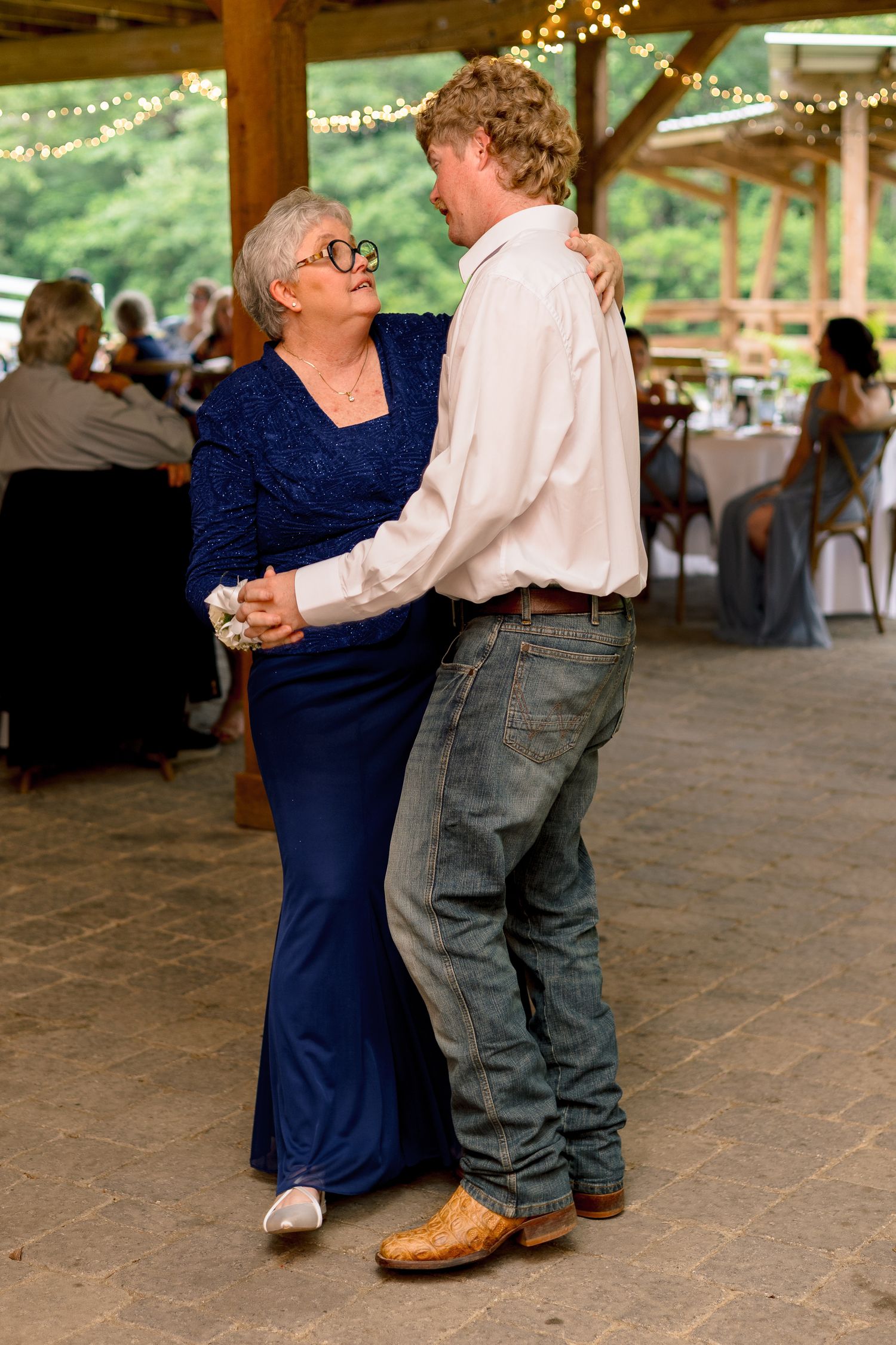 Groom in white shirt and jeans shares mother-son dance under string lights at rustic outdoor wedding reception.