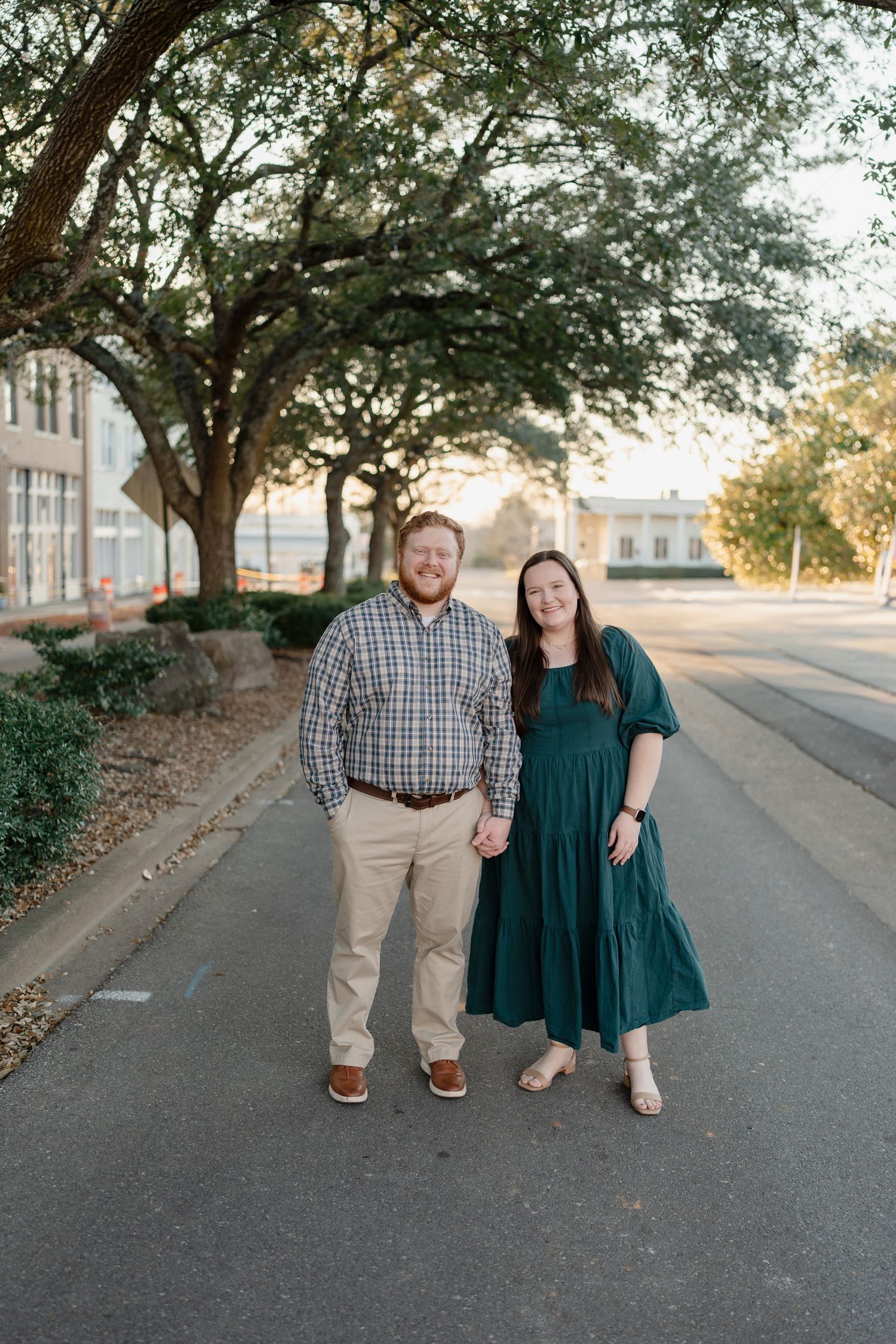 Smiling couple holds hands on tree-lined street, woman in emerald maxi dress and man in plaid shirt.