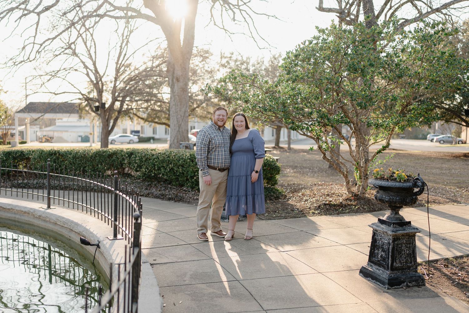 Couple smiling together by a garden fountain with lush greenery and warm sunlight in an outdoor park setting.