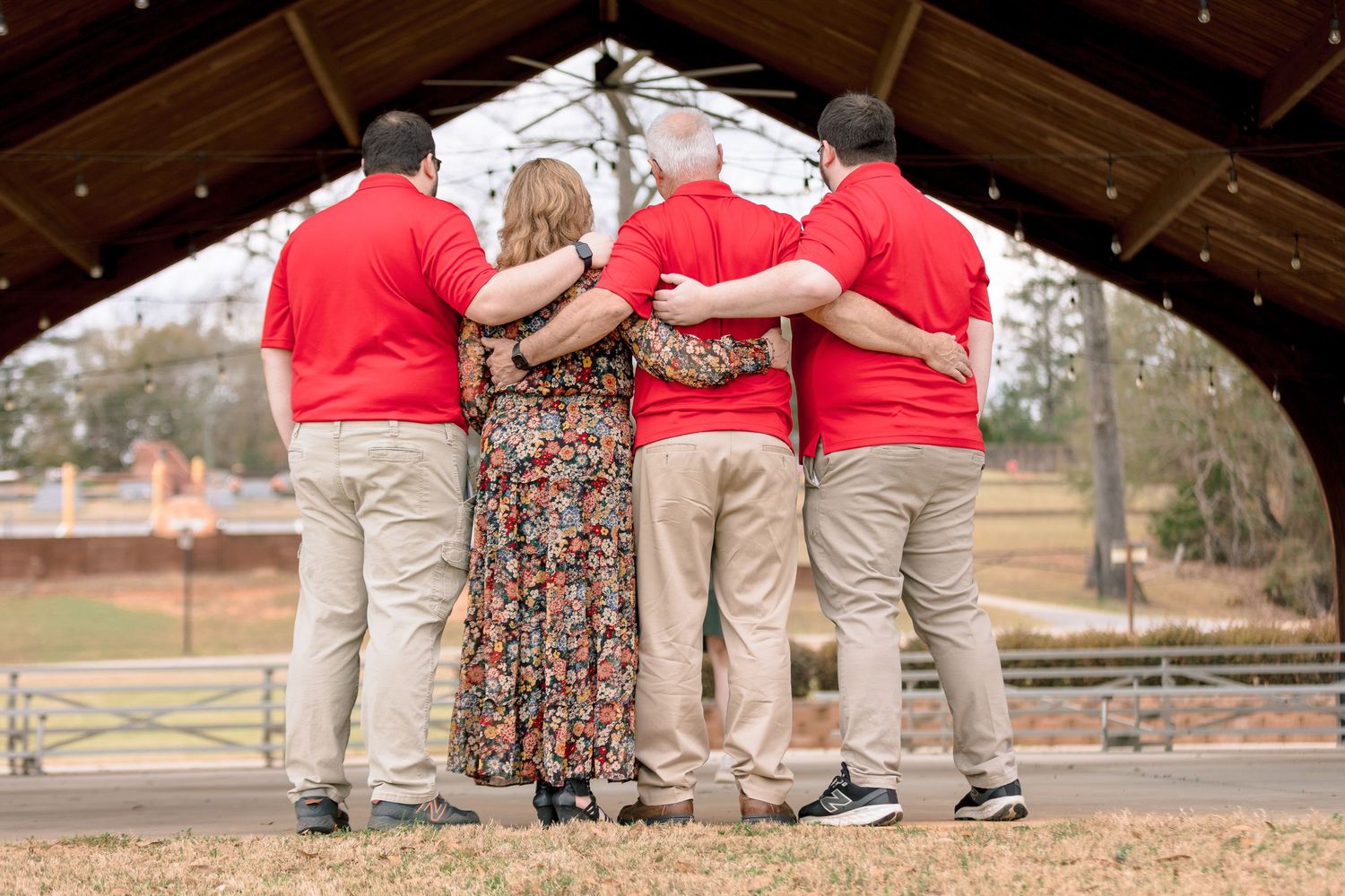 Family group hugging under a wooden pavilion, viewed from behind, wearing matching red shirts.