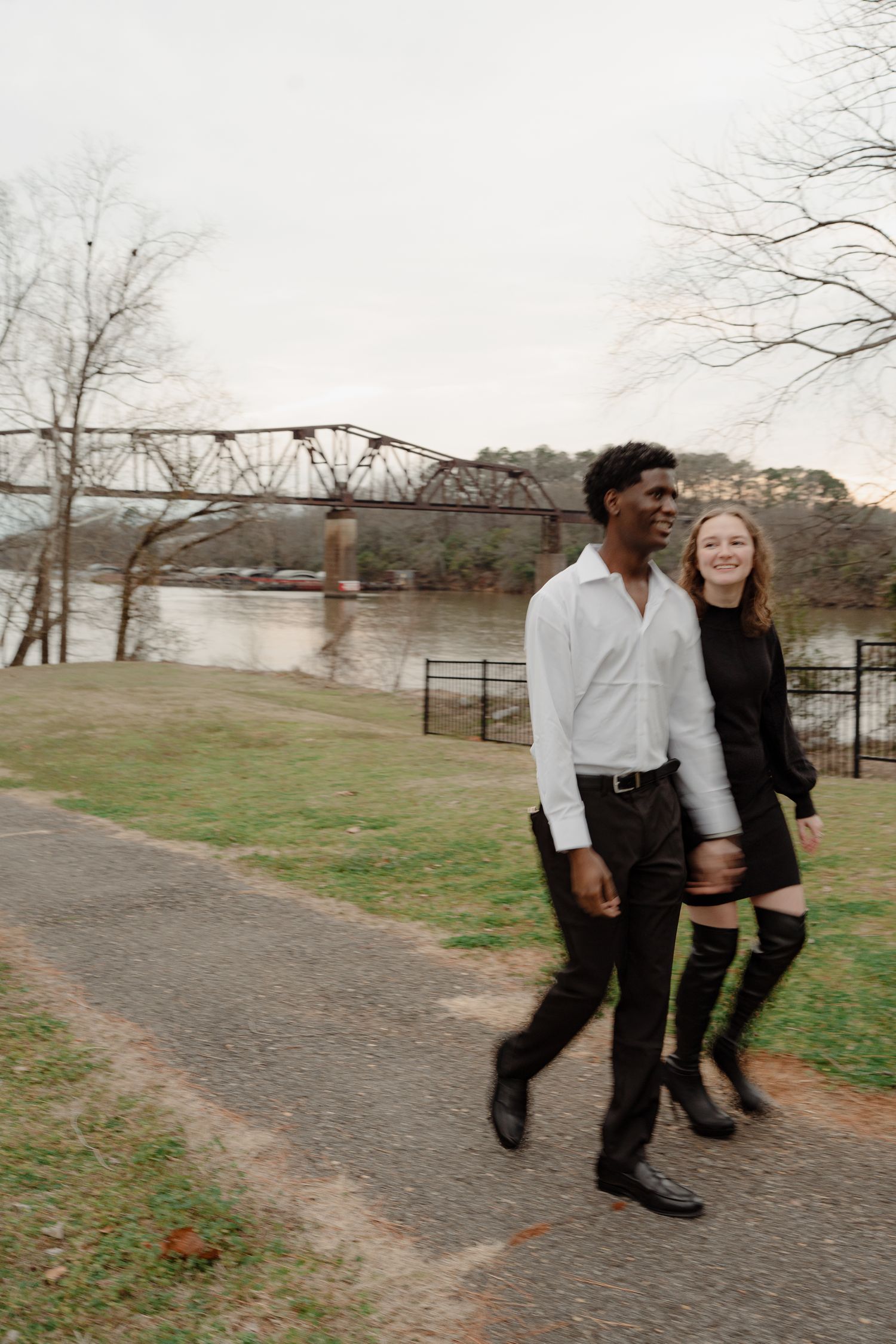 A smiling interracial couple walks hand-in-hand along a park path with a bridge backdrop.