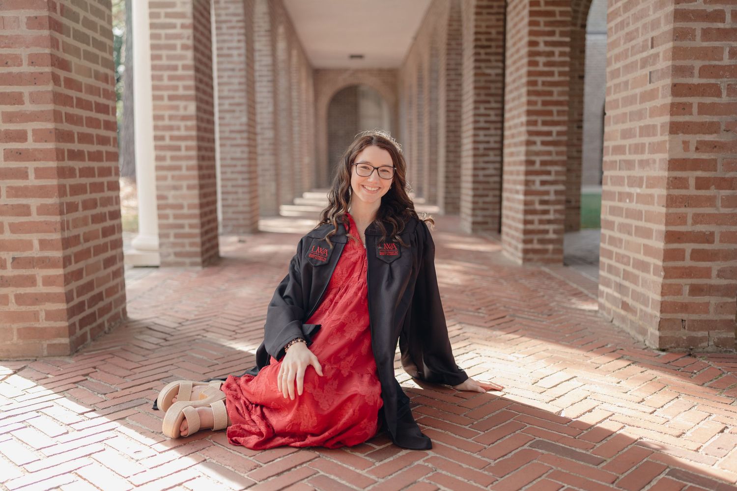 A person in a red dress and black jacket sits on brick steps in a covered outdoor corridor with arches.