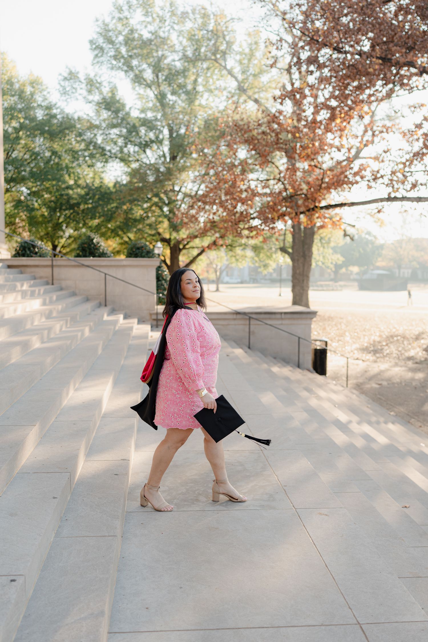 Graduate posing on concrete steps in pink dress holding graduation cap against autumn backdrop.
