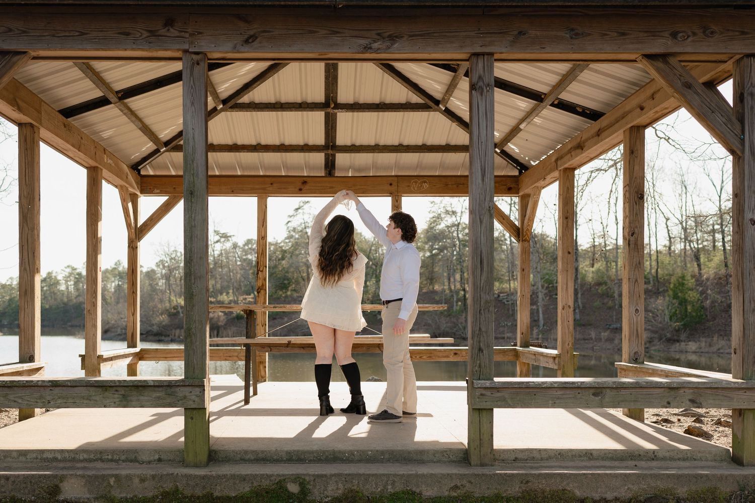 Couple dancing together under a rustic wooden pavilion overlooking a serene lake in a wooded setting.
