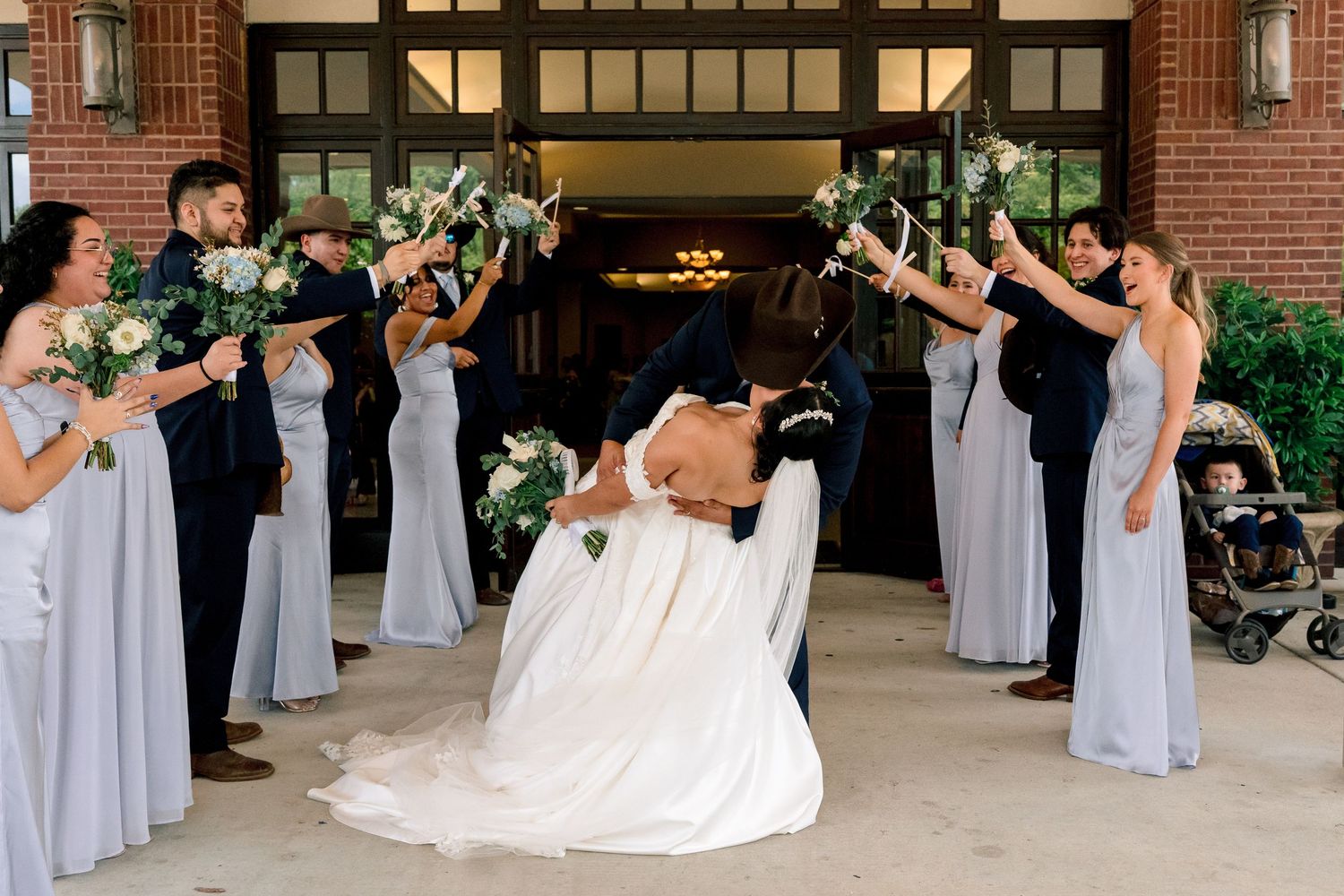 Wedding party in light gray dresses poses with bride in white gown outside brick building.