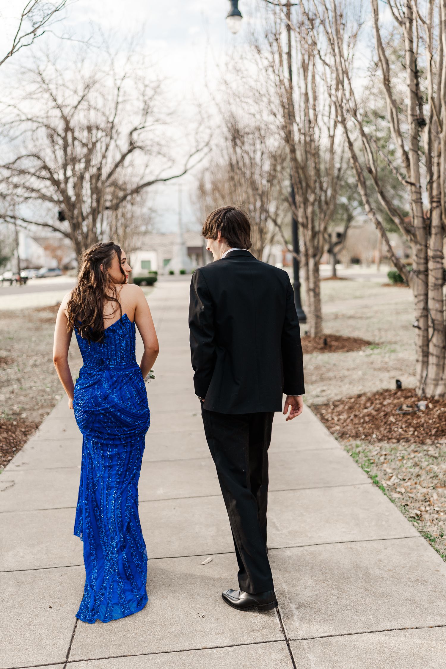 Couple in formal wear walking on sidewalk path surrounded by bare winter trees.