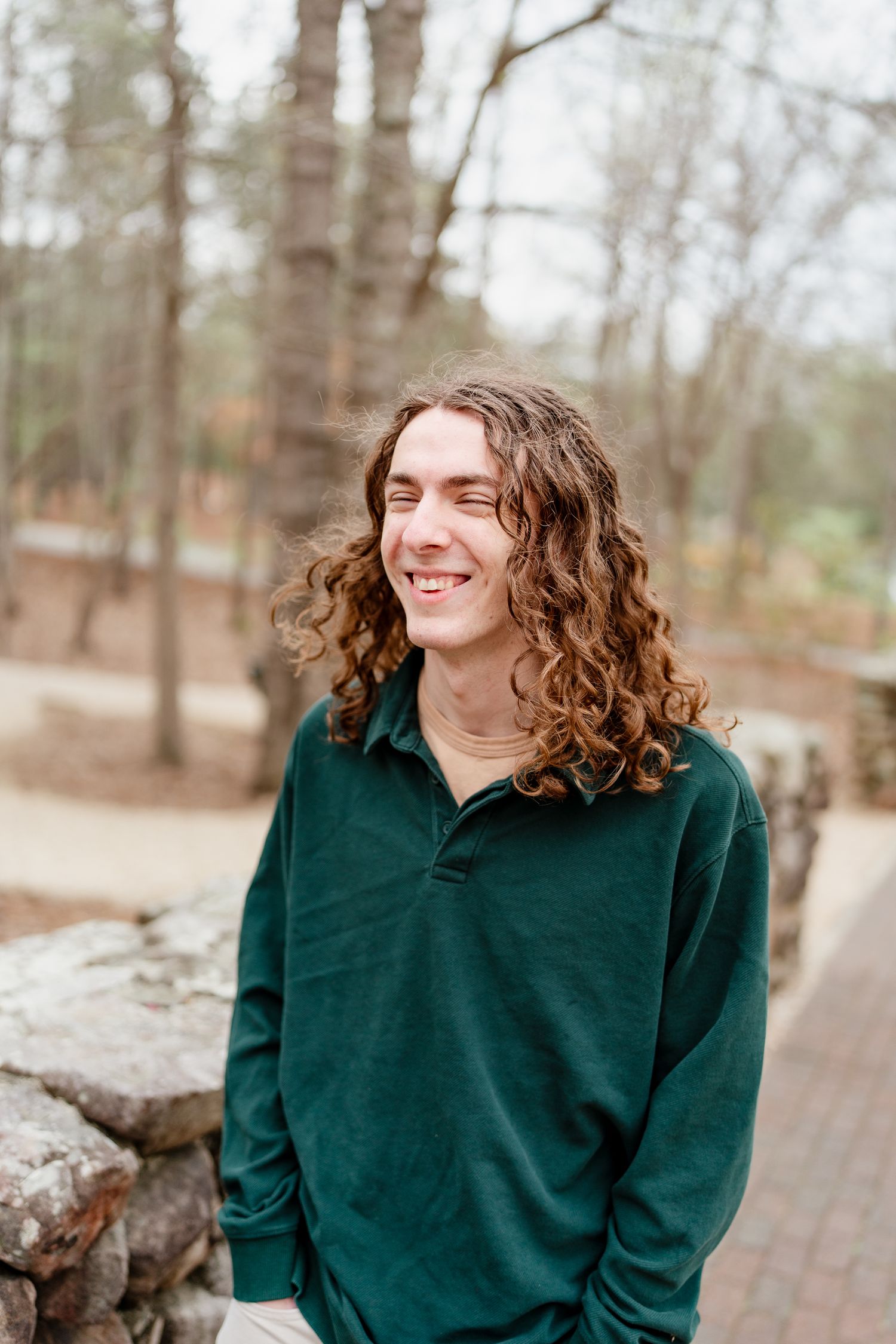 Casual portraits by a stone wall in a forest setting wearing a dark green sweatshirt.