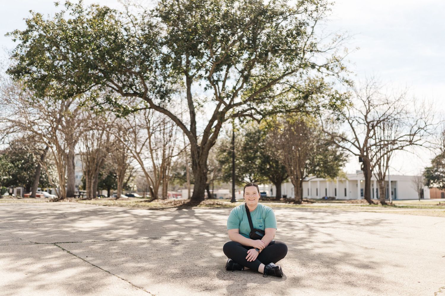 Person crouching in empty courtyard surrounded by trees on sunny day.