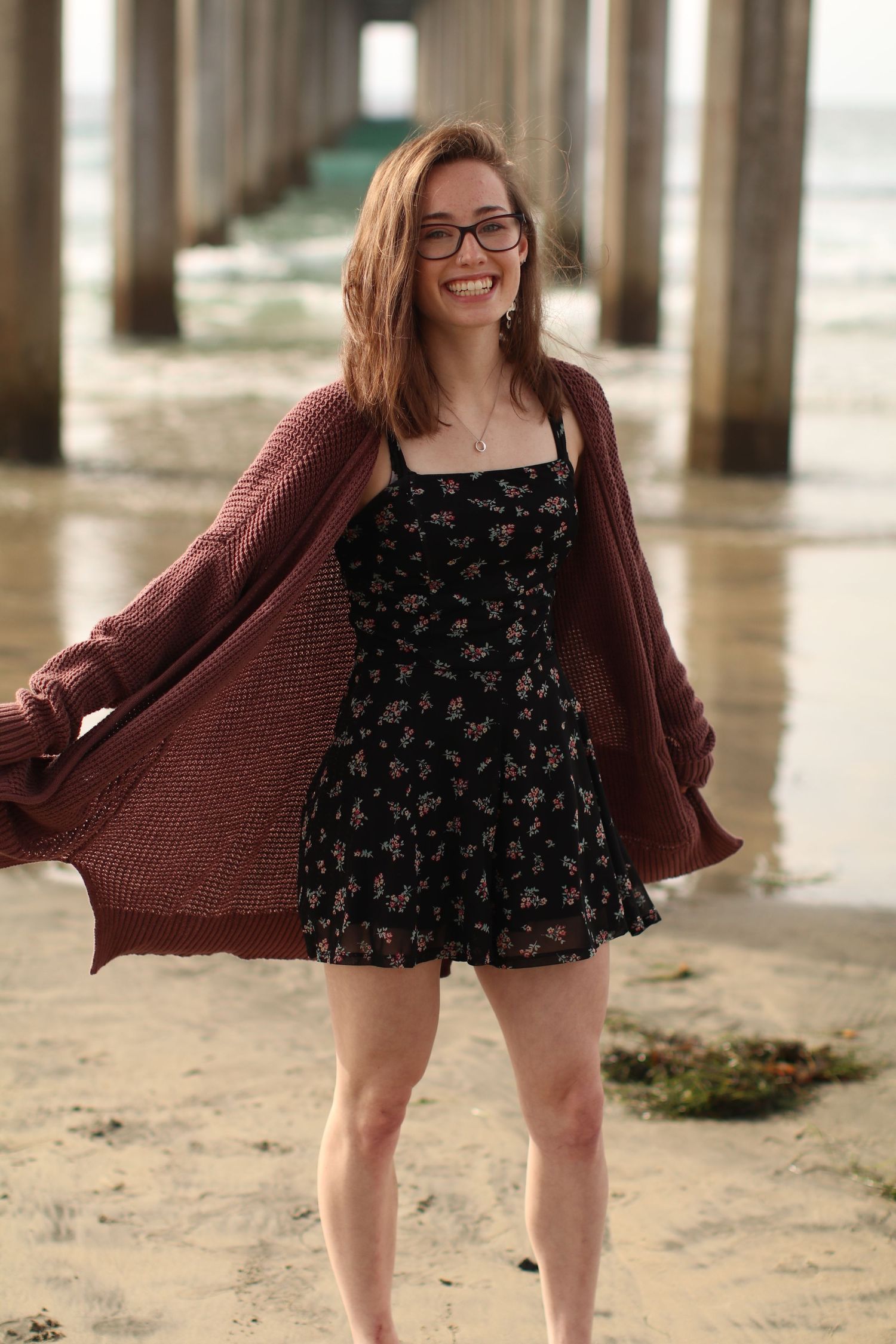 Playful beachside photo under pier wearing floral dress and oversized knit cardigan.
