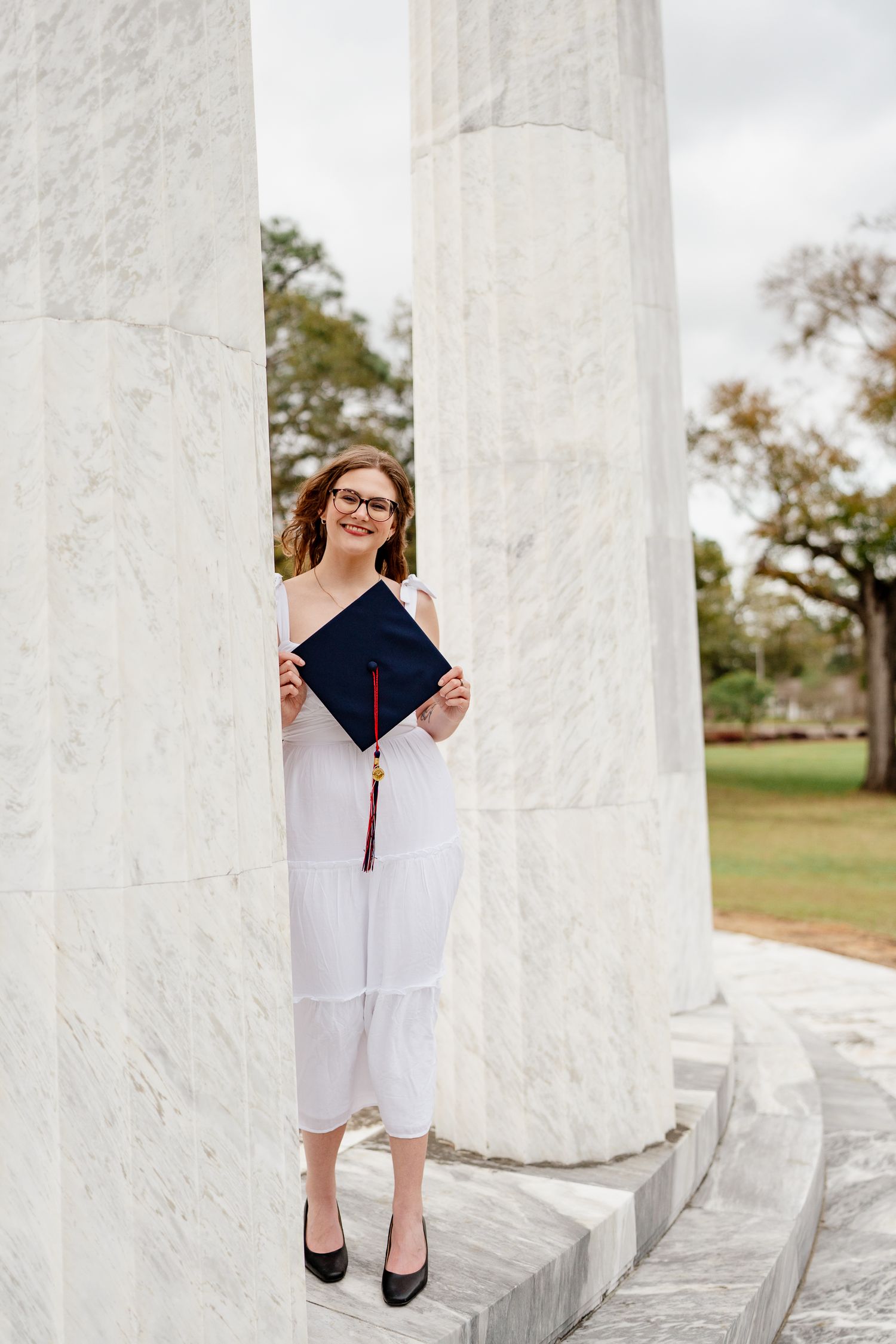 Graduate poses playfully between white marble columns in graduation attire.