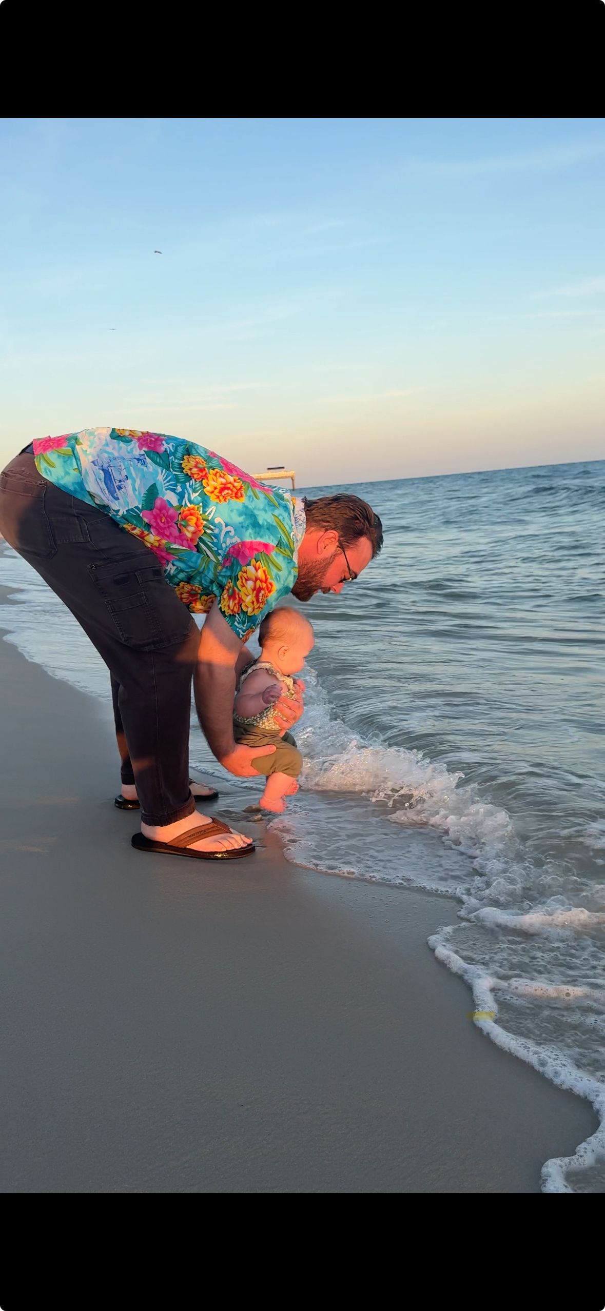 Person in colorful clothing bends down to touch ocean waves at sunset on a beach.