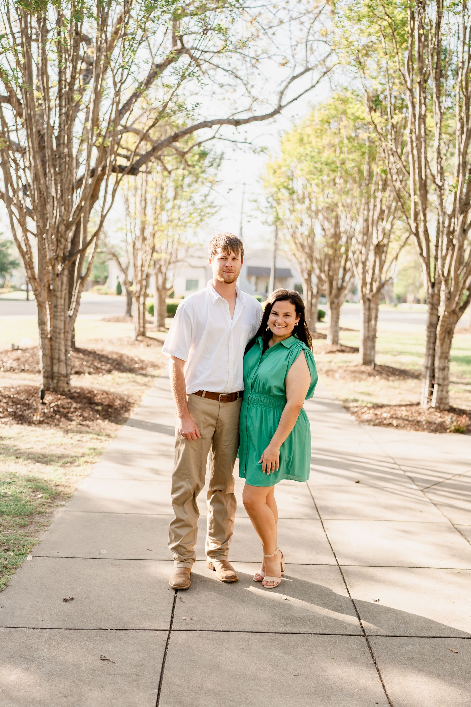 Couple in green dress and white shirt pose together on tree-lined pathway.