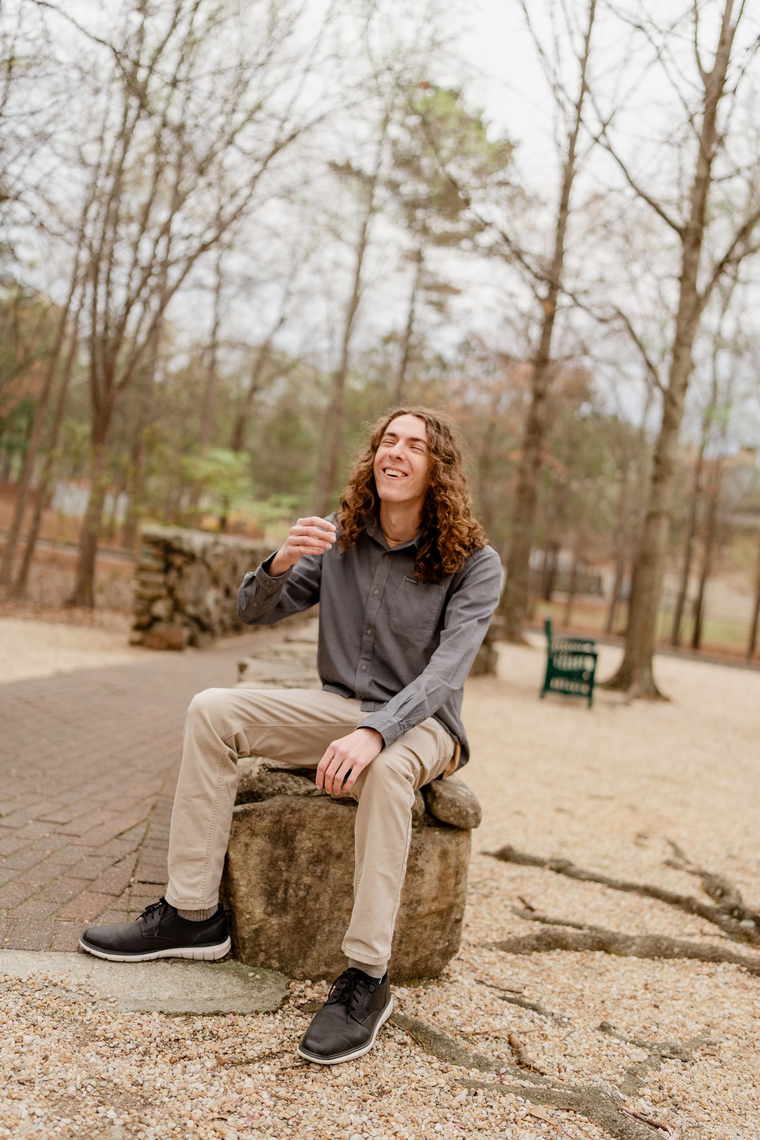 Person sitting casually on a tree stump in a wooded park area wearing khakis and a gray sweater.