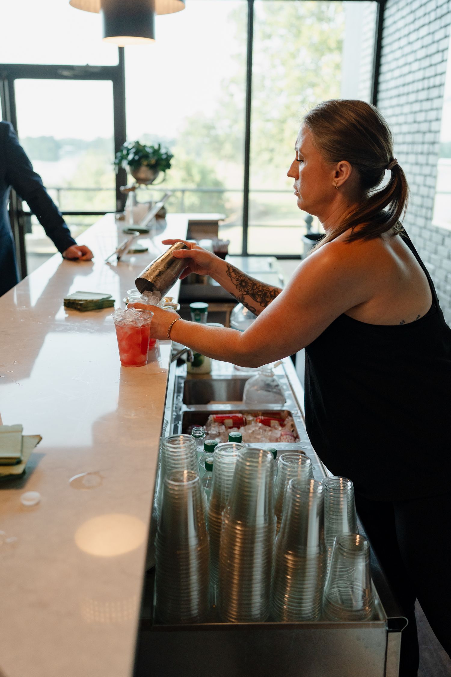 A bartender in black prepares drinks at a modern bar station with stacked cups and beverages.