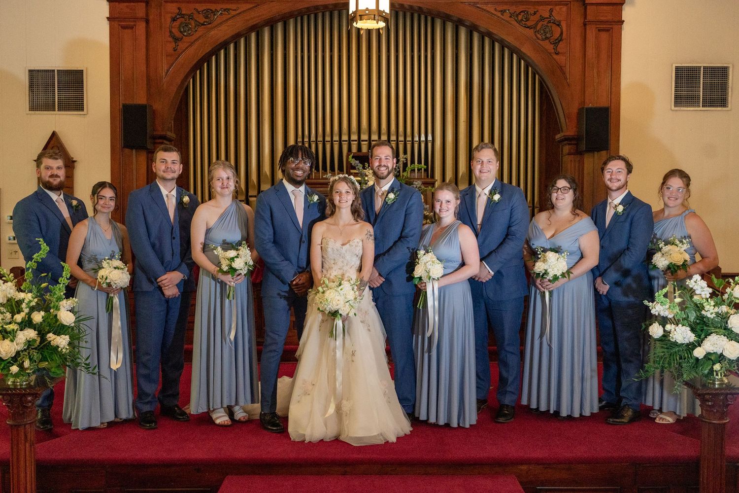Wedding party poses formally in a church sanctuary with decorative organ pipes and floral arrangements.