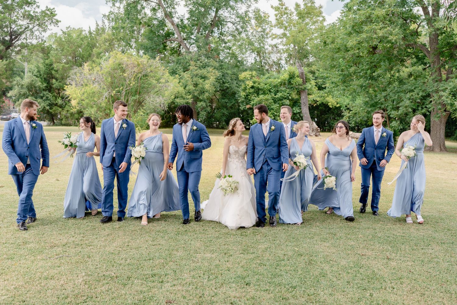 Wedding party in coordinating navy and light blue attire poses together on grassy lawn during outdoor celebration.