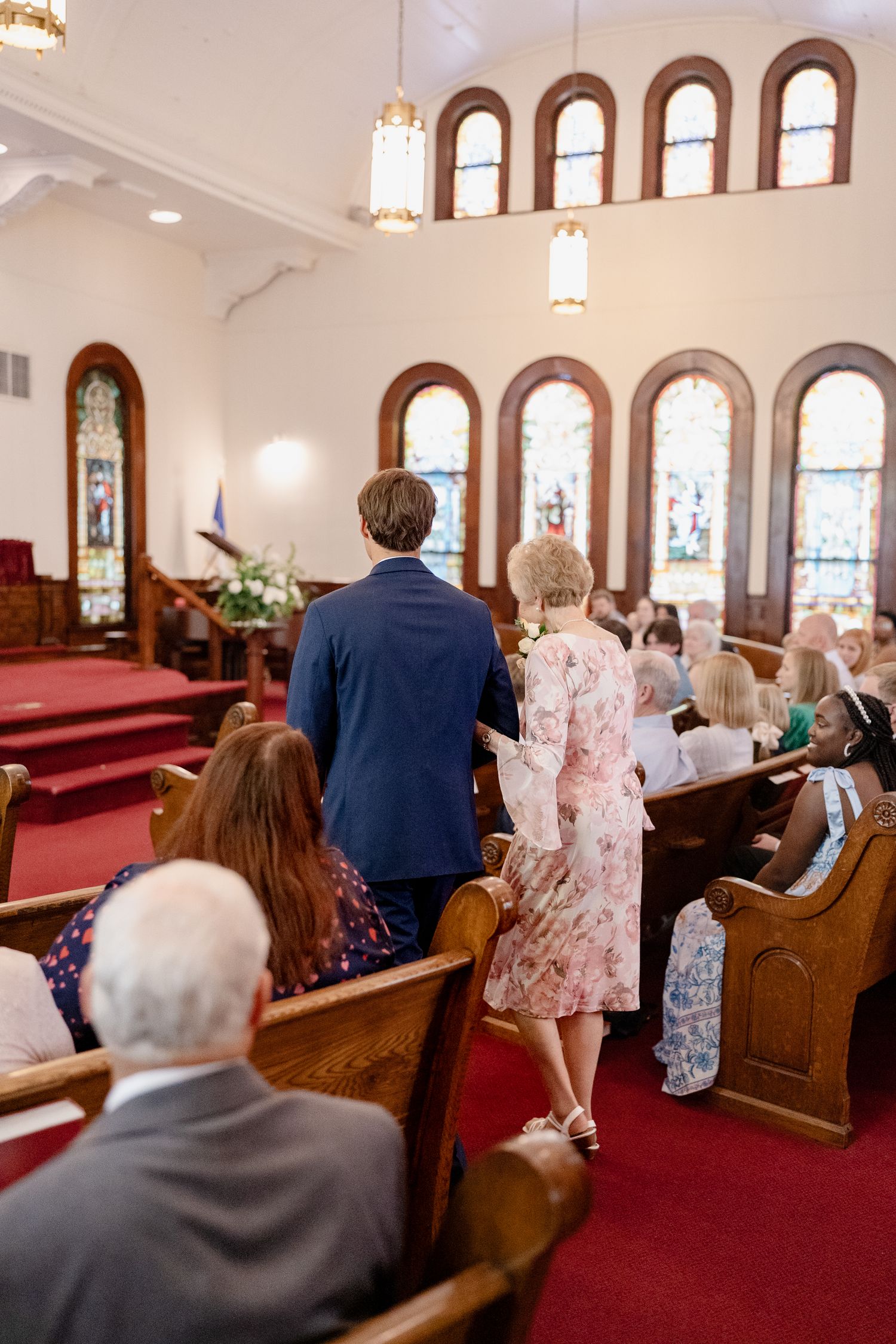 Wedding guests seated in wooden pews of a traditional church with stained glass windows and red carpet.
