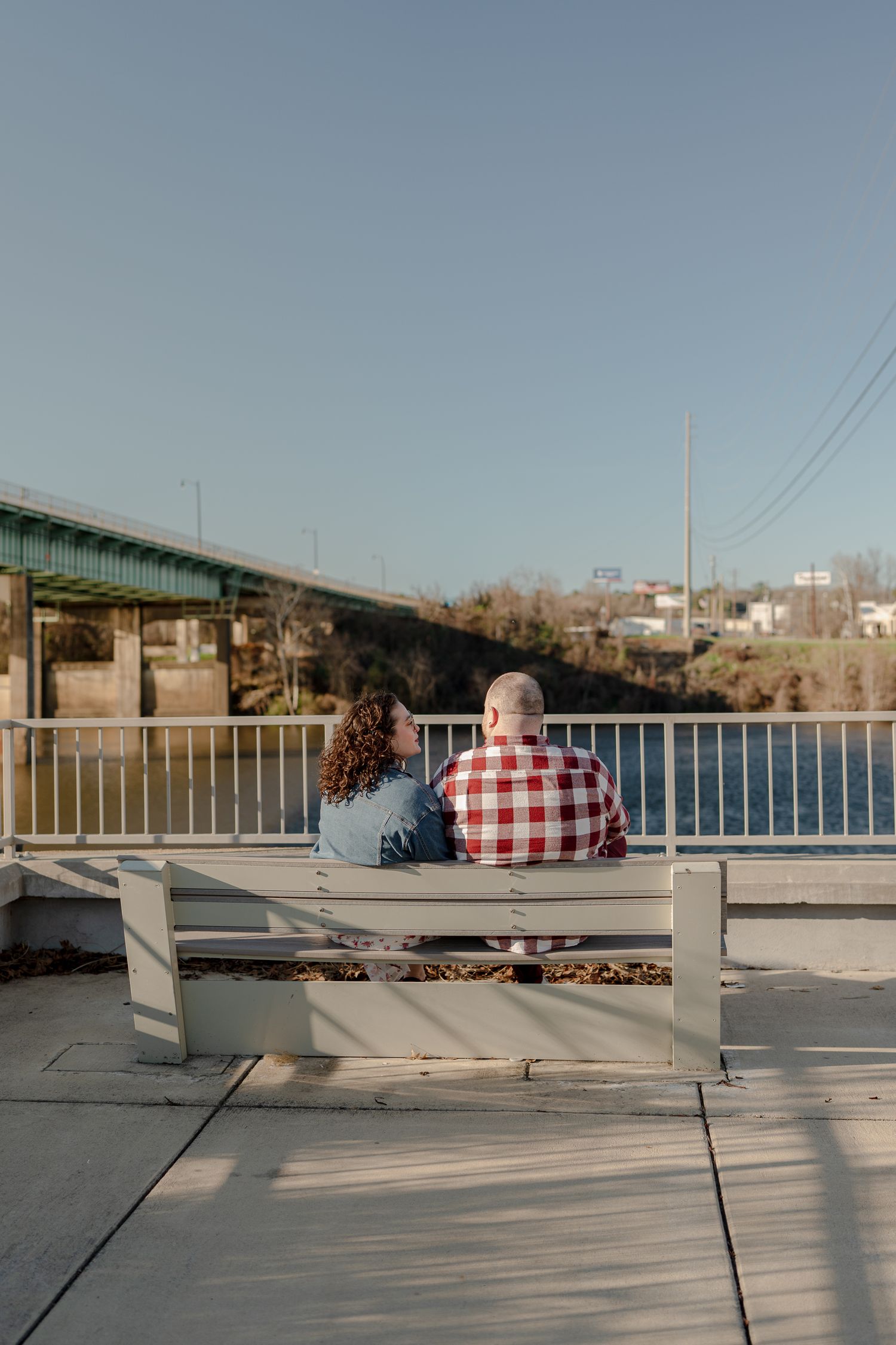 A couple sits together on a concrete ledge overlooking a waterfront on a sunny afternoon.