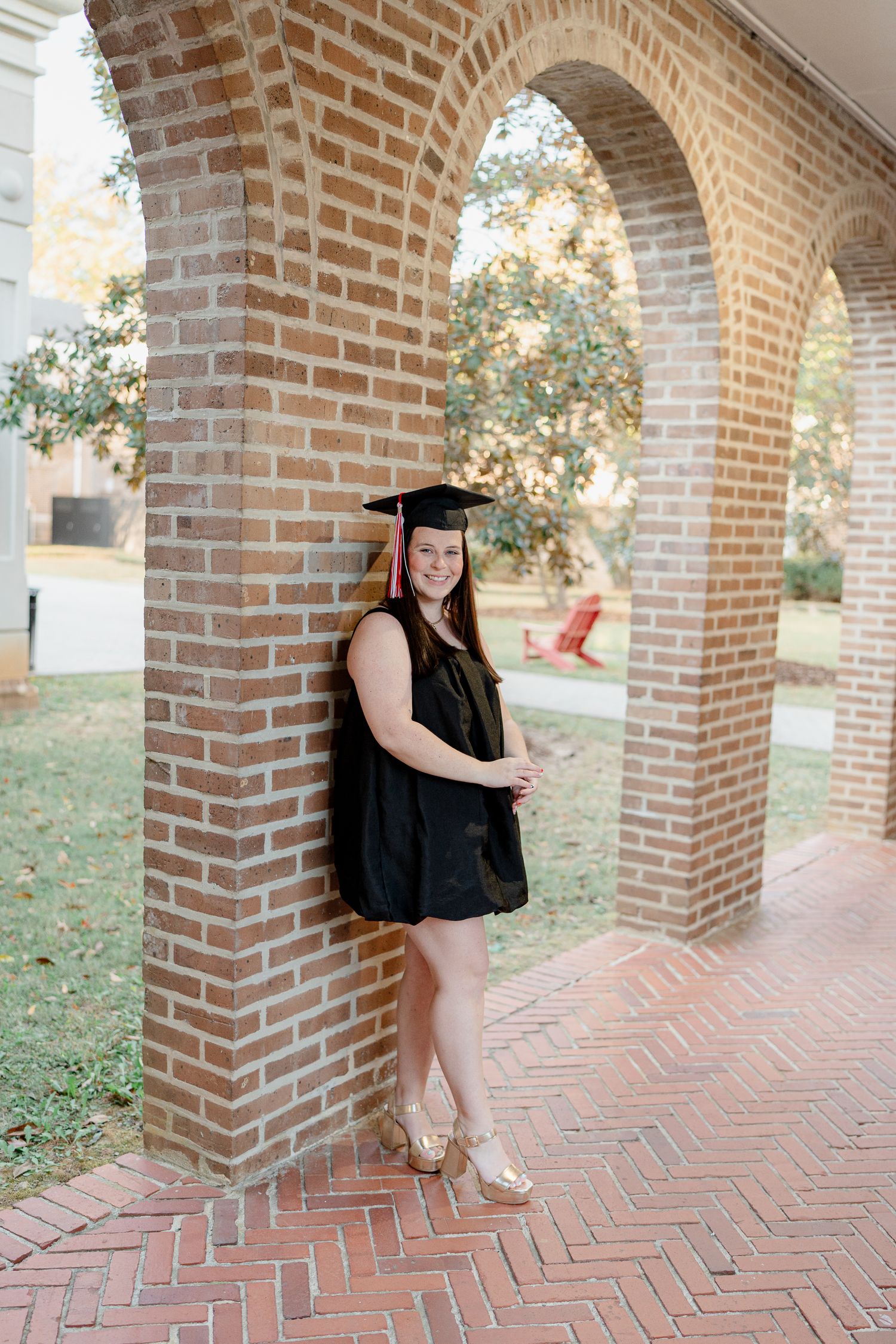 Graduate in black cap and gown poses under brick archways on a patterned brick walkway.
