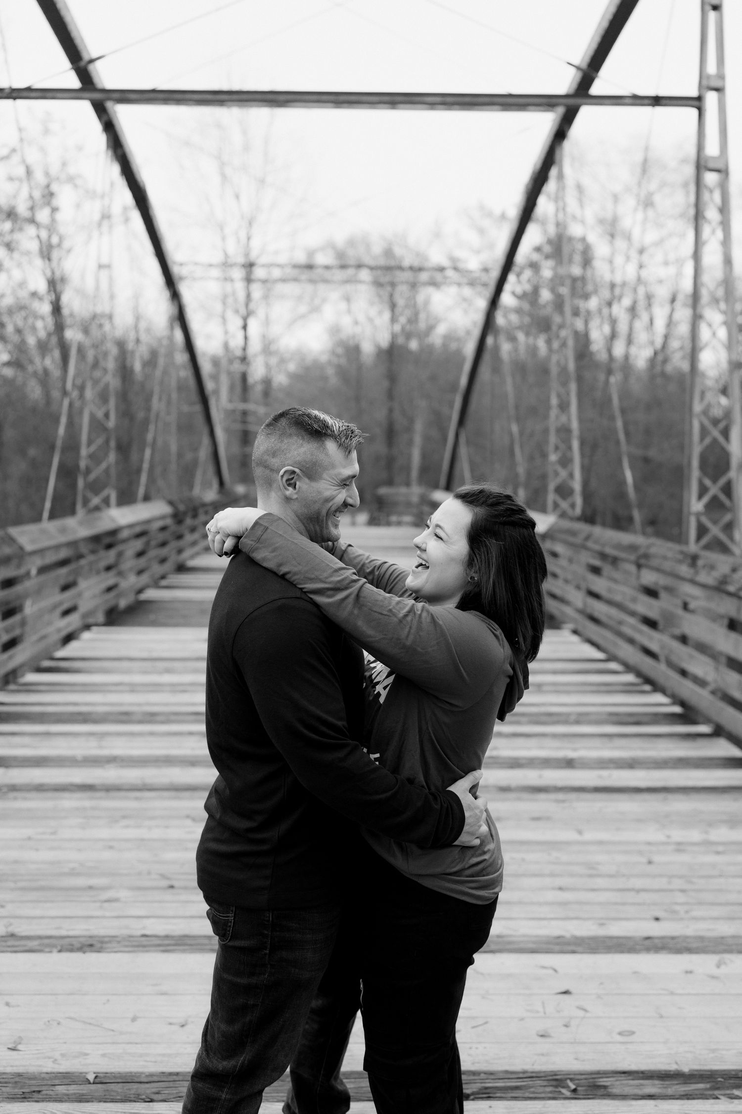 A romantic couple embraces on a wooden bridge surrounded by bare winter trees.