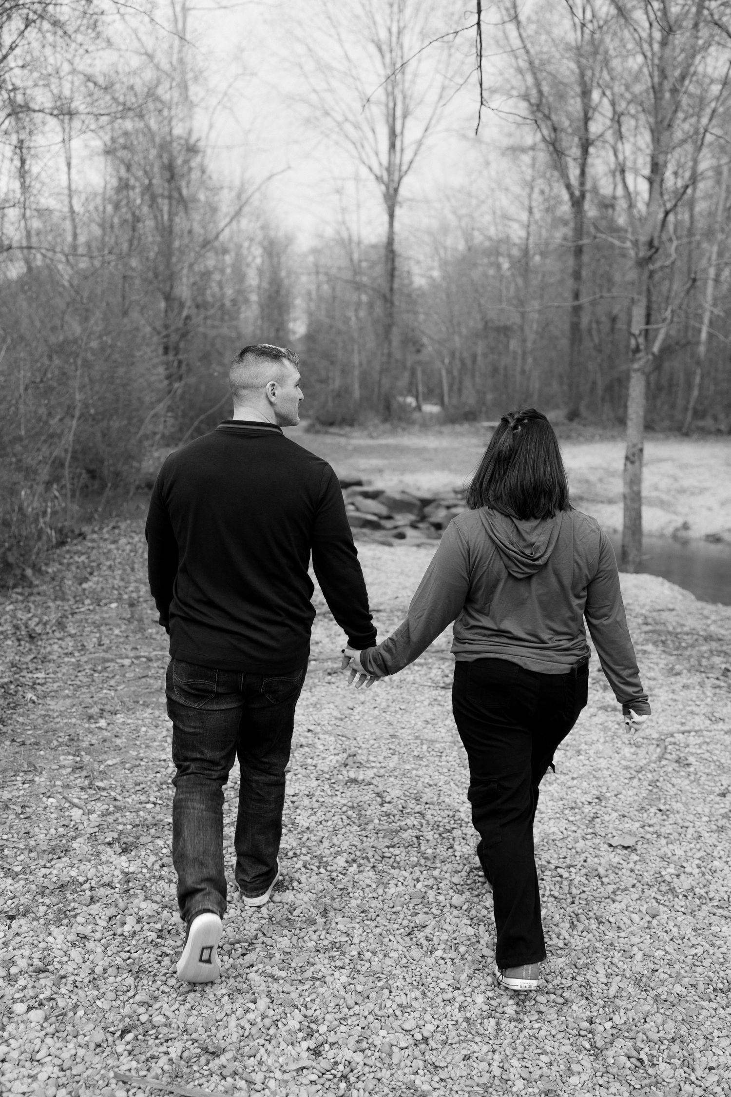 A couple walks hand in hand along a gravel path through a wooded area in black and white.