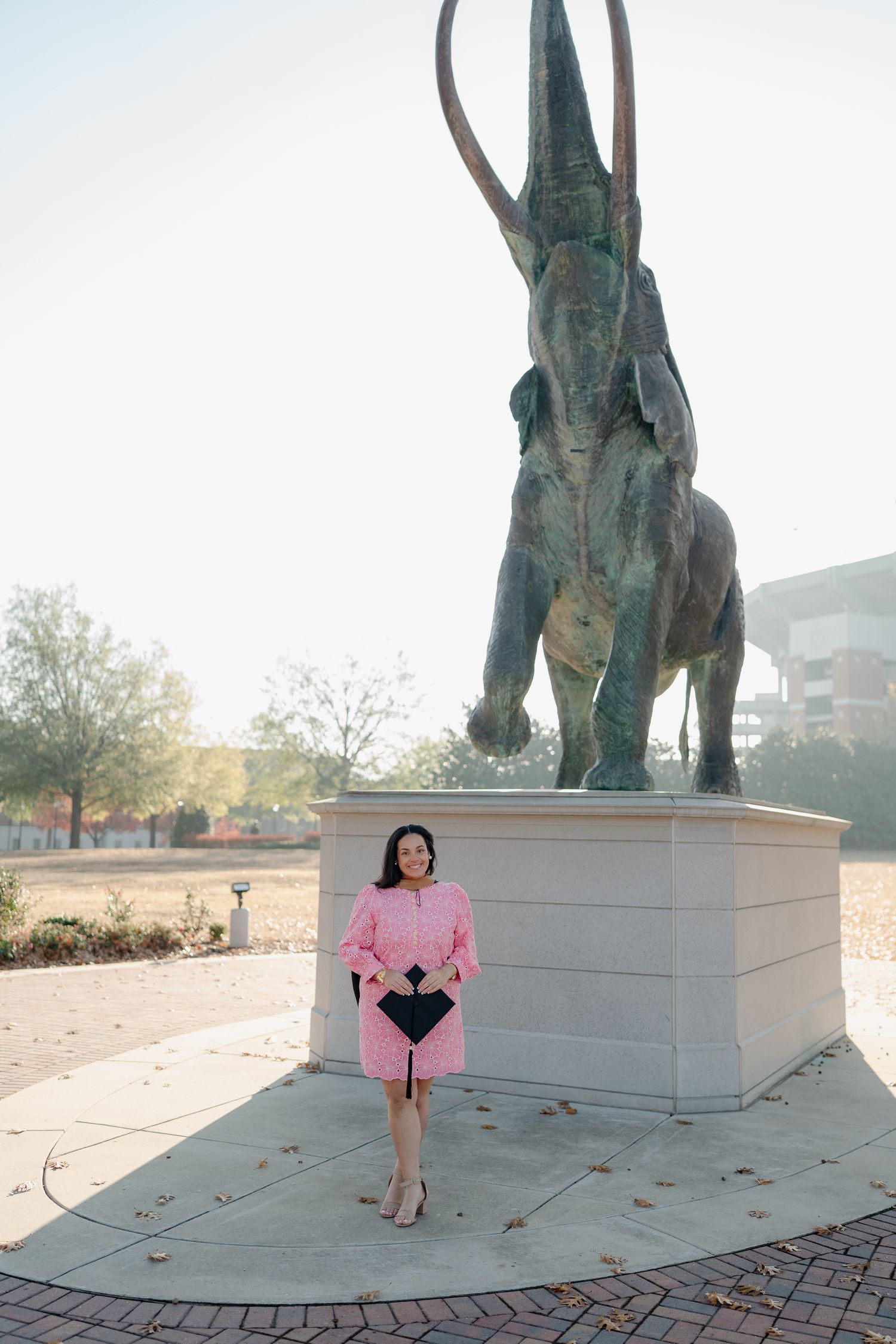 Graduate in pink outfit poses in front of large bronze elephant statue on campus plaza.