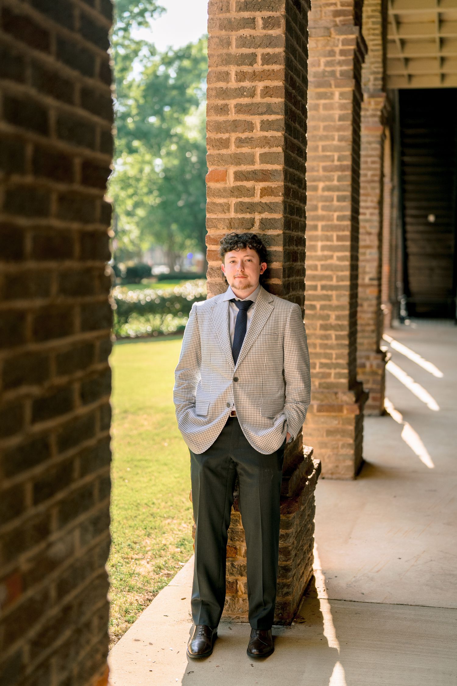 Young male graduate in grey blazer and tie poses between brick columns on University of Alabama campus.