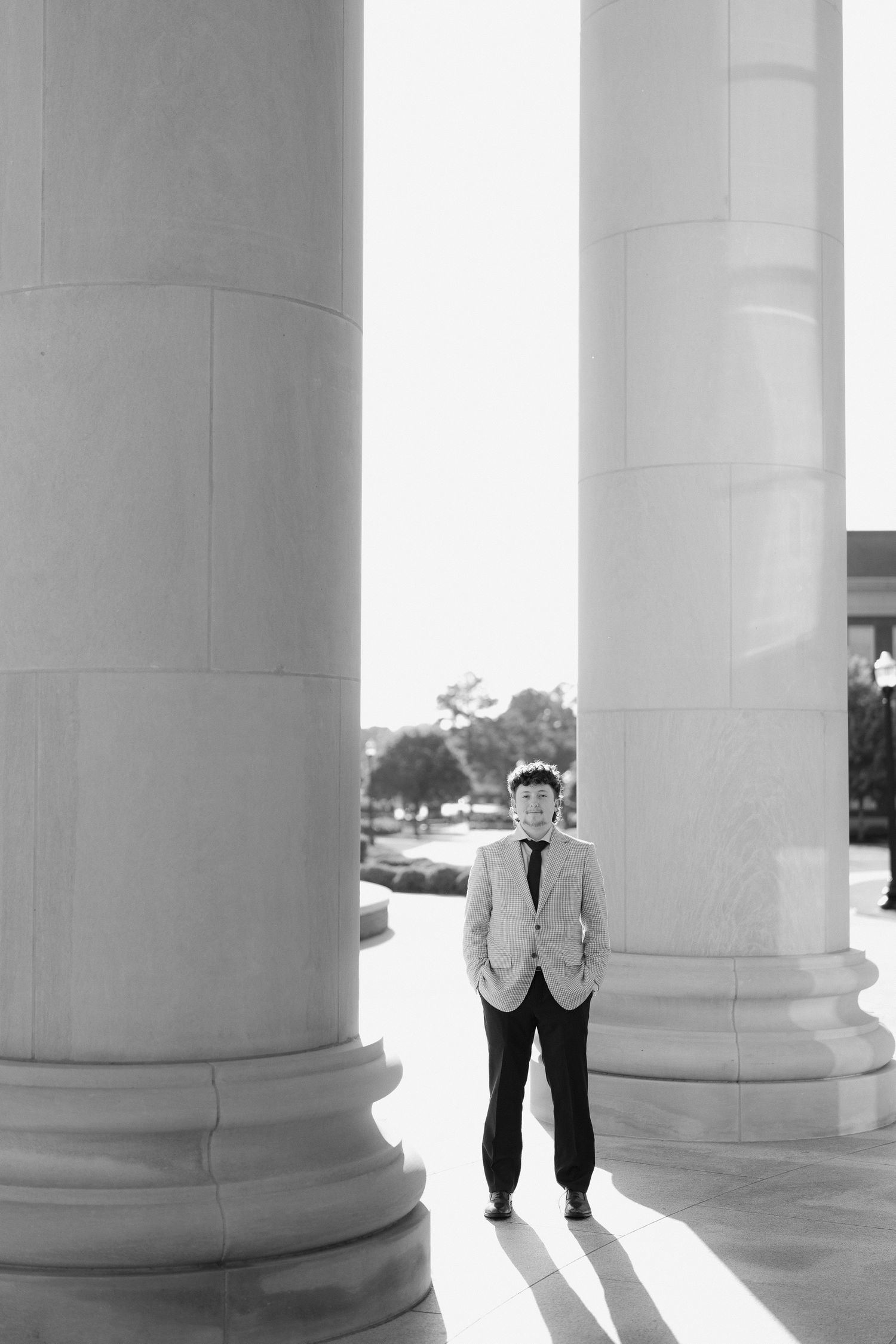 Black and white photo of young man in suit standing between two large white classical columns in bright light.