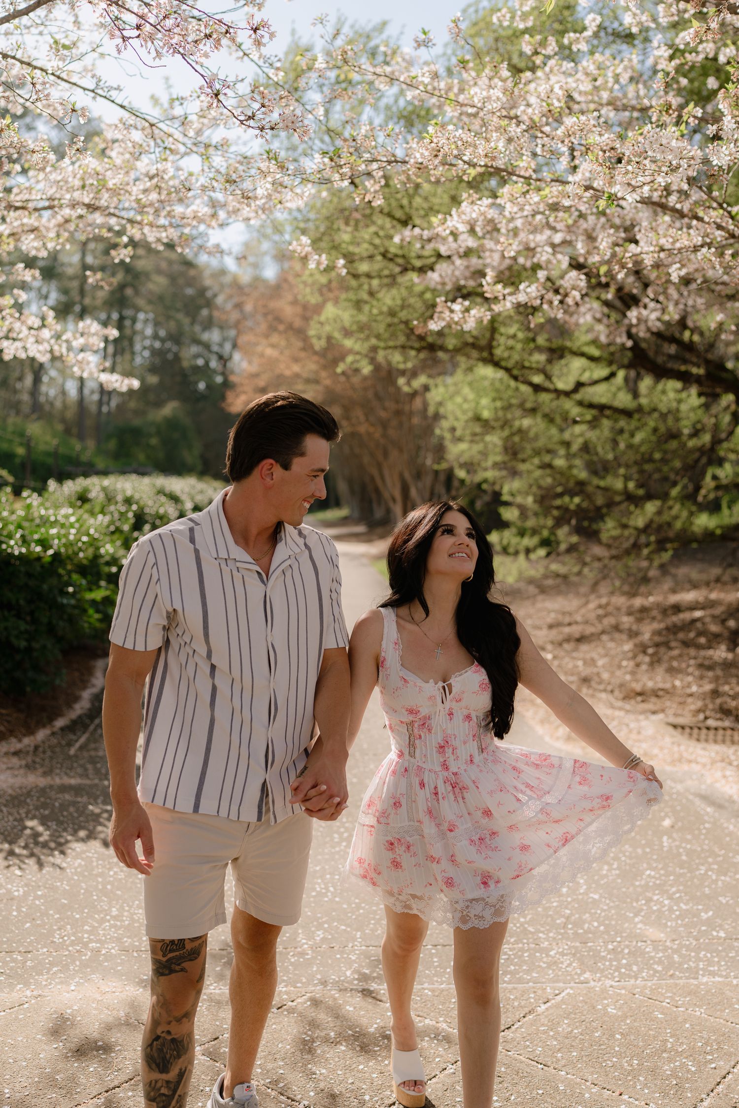 Happy couple holding hands walking under white flowering cherry blossom trees on sunny garden path.