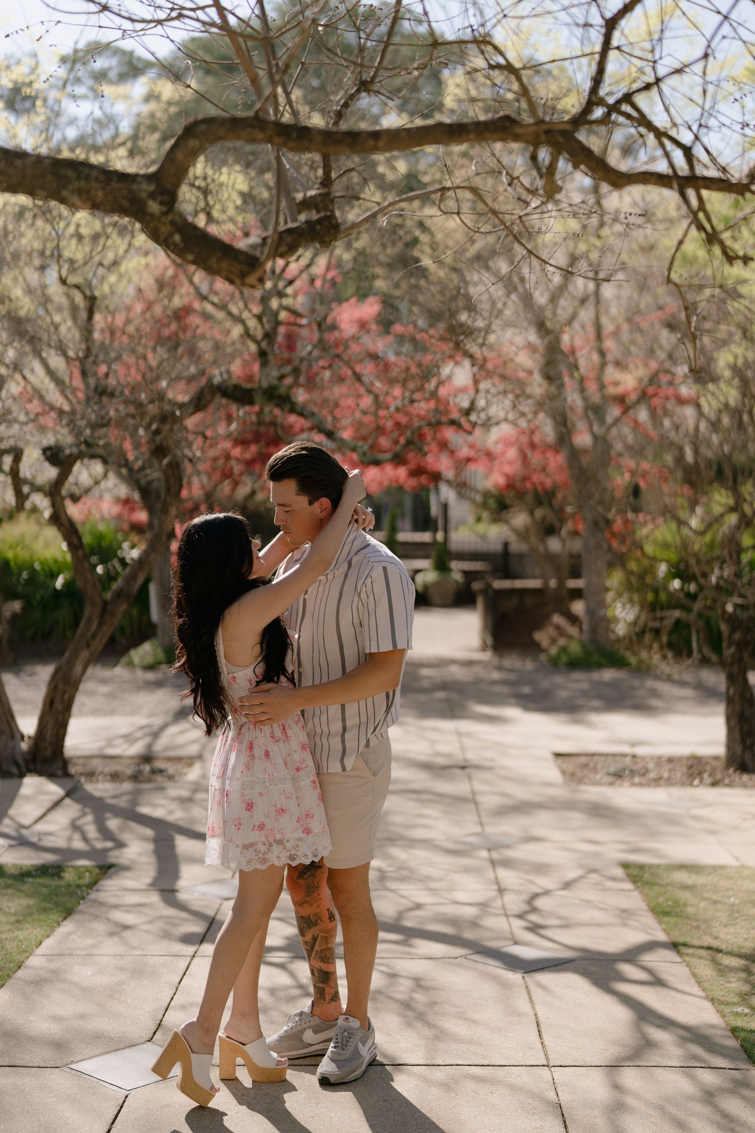 Couple embracing romantically on a sunny path surrounded by blooming pink trees in a garden setting.