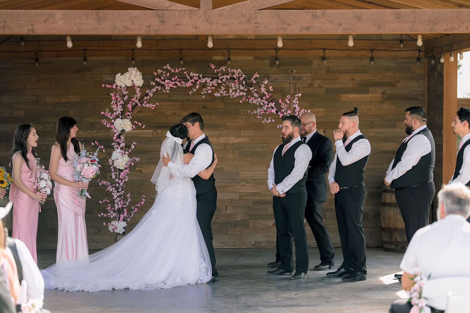Bride and groom share a kiss at the altar beneath a floral cherry blossom arch at a rustic barn wedding.