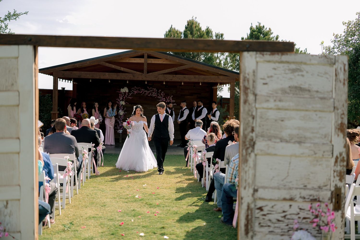 Bride and groom walk down outdoor wedding aisle framed by rustic doors with guests seated on white chairs.