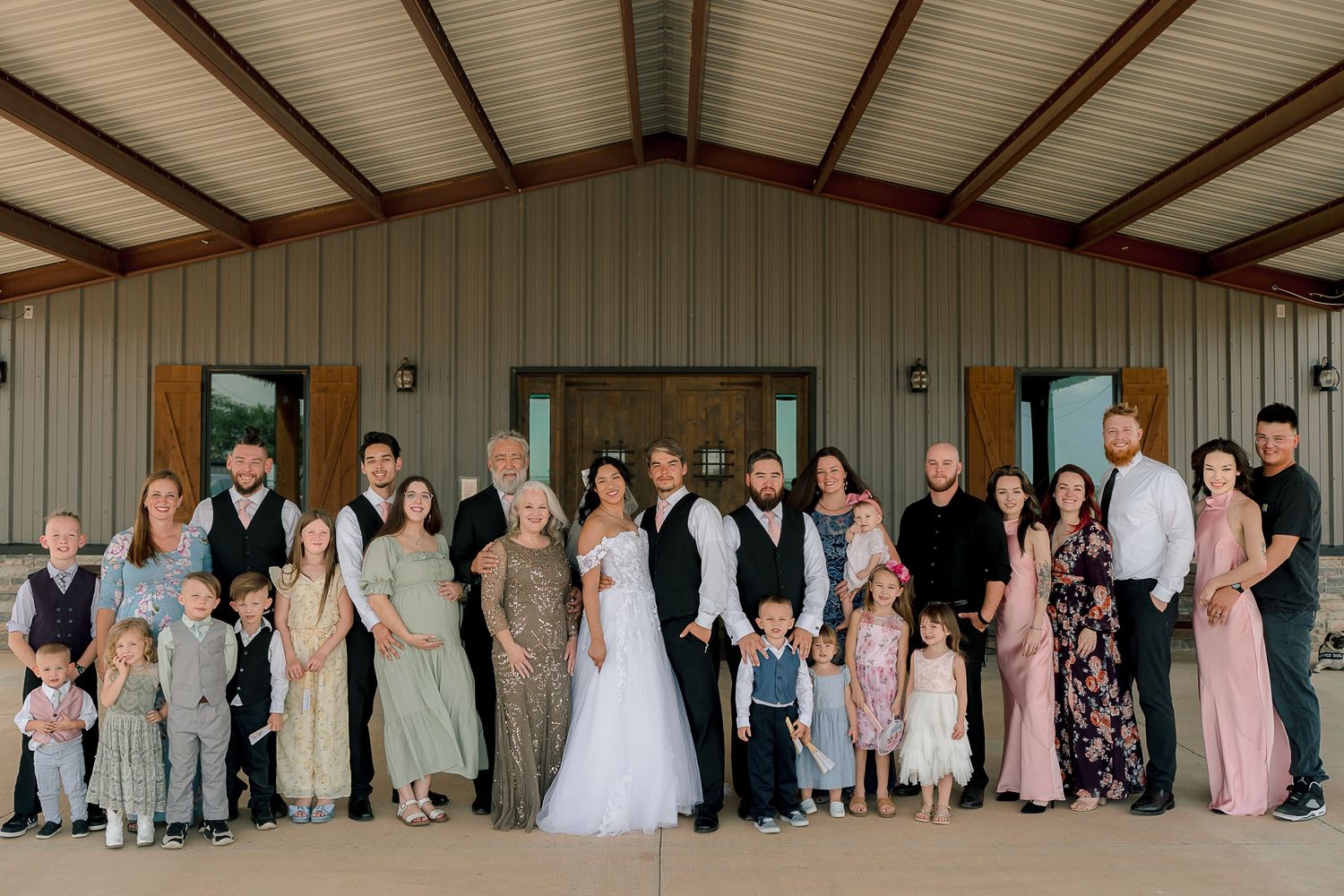 Large wedding family group portrait posed outside a rustic barn venue on a sunny day.
