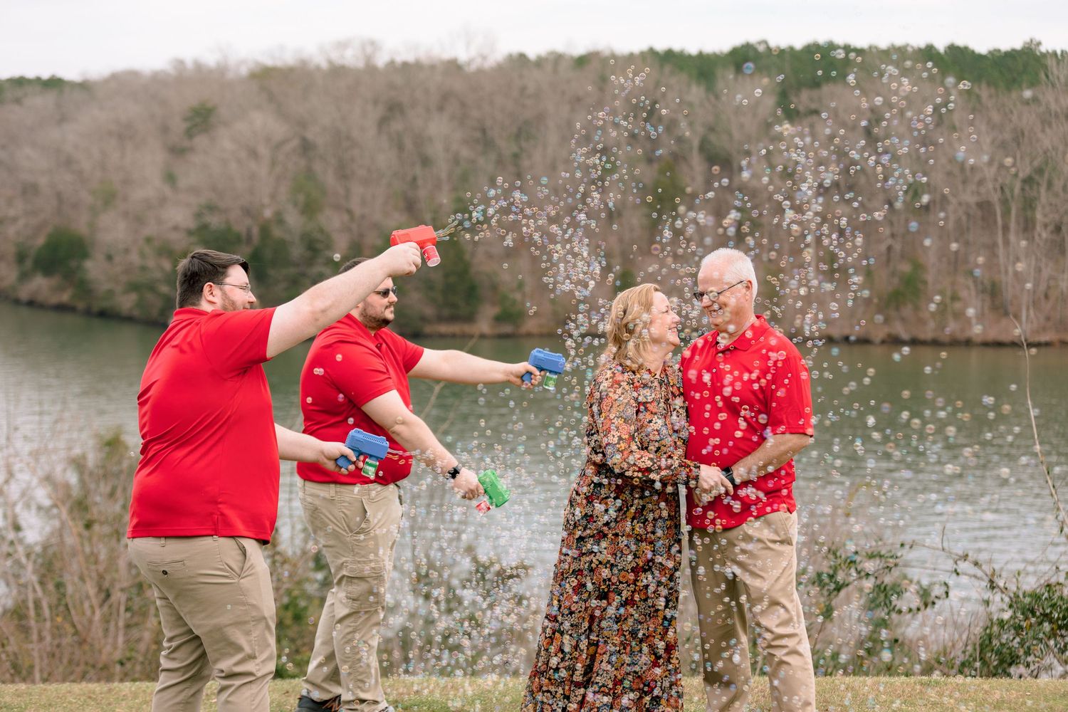 Family celebrates with confetti poppers outdoors by a lake after a wedding ceremony.