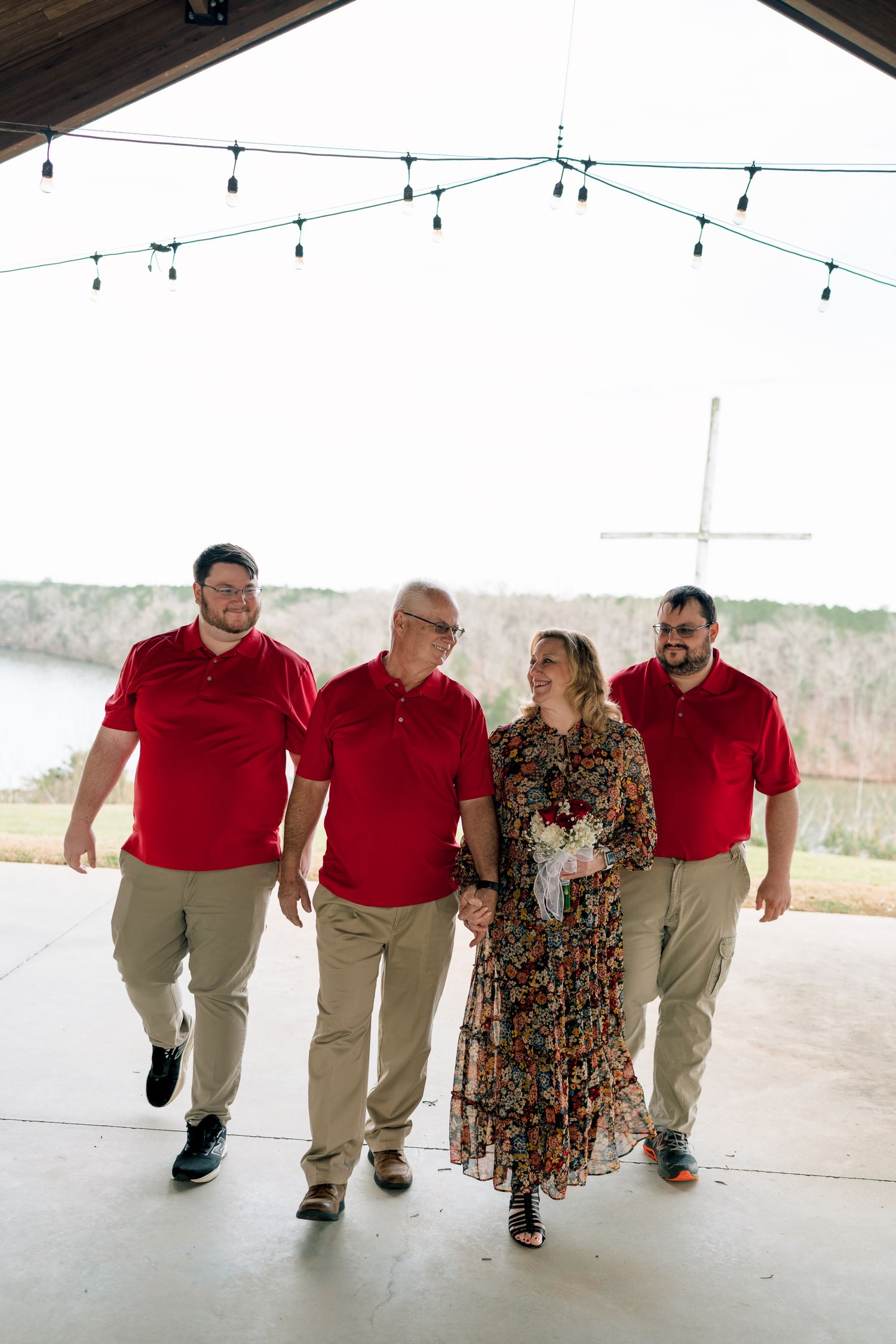 Family of four in red and floral attire walking under string lights near a lakeside pavilion.