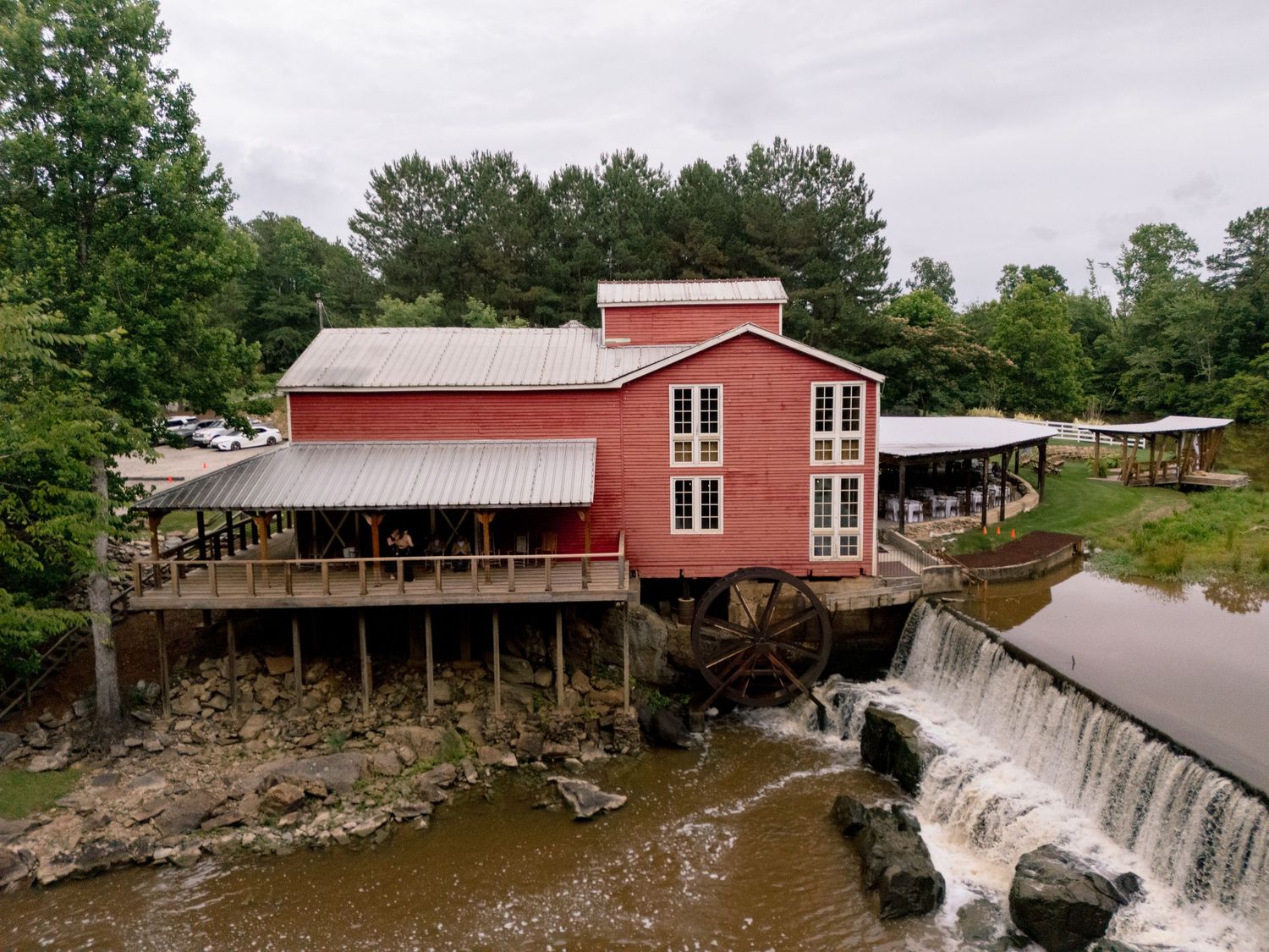 Aerial view of red historic grist mill with water wheel and waterfall on scenic wooded river.