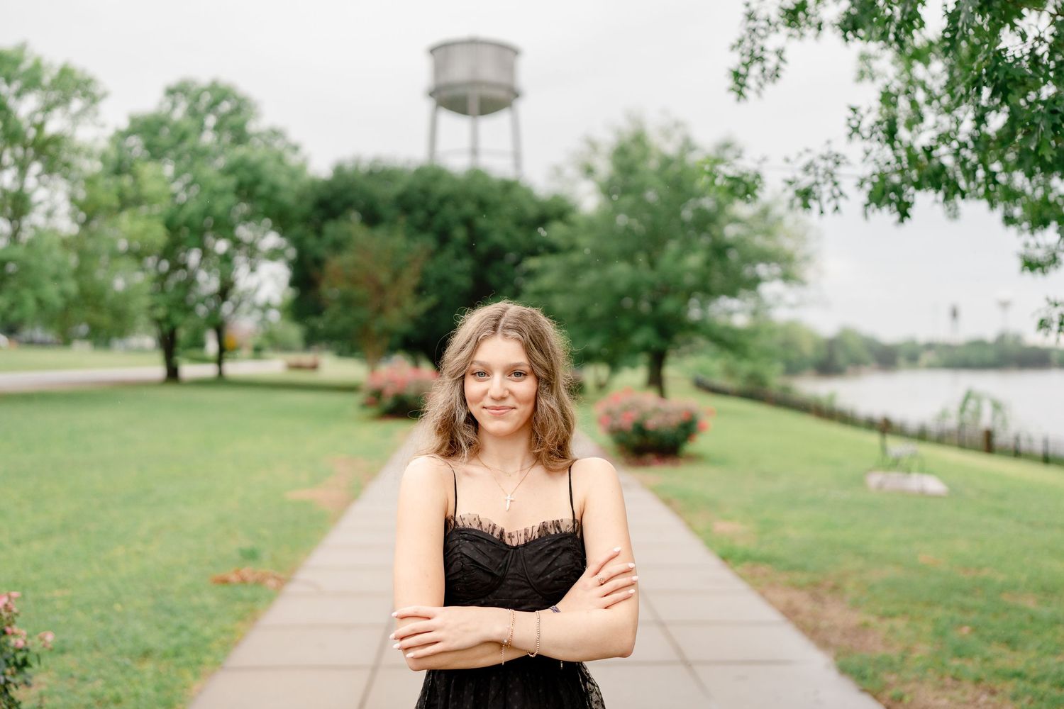 A person stands outdoors on a paved path with a water tower visible in the background among green trees.