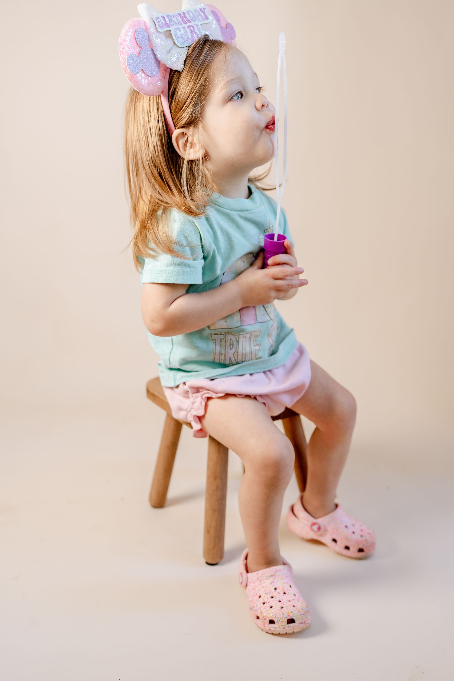 Pink Crocs and a mint colored shirt appear against a minimalist beige studio setting.