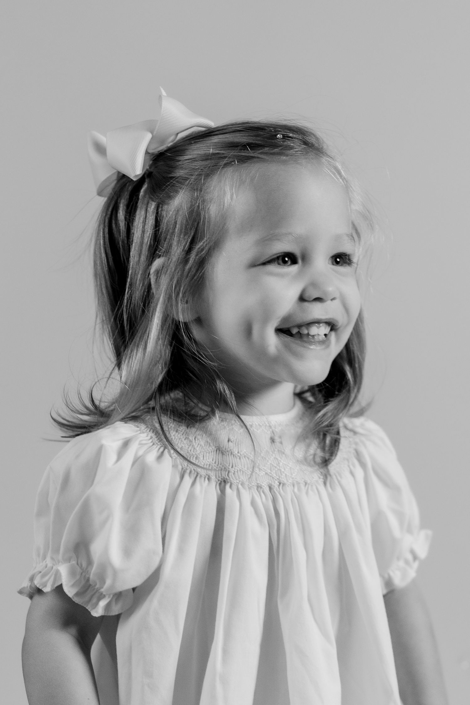 Child wearing white ruffled dress poses playfully against grey background in portrait series.