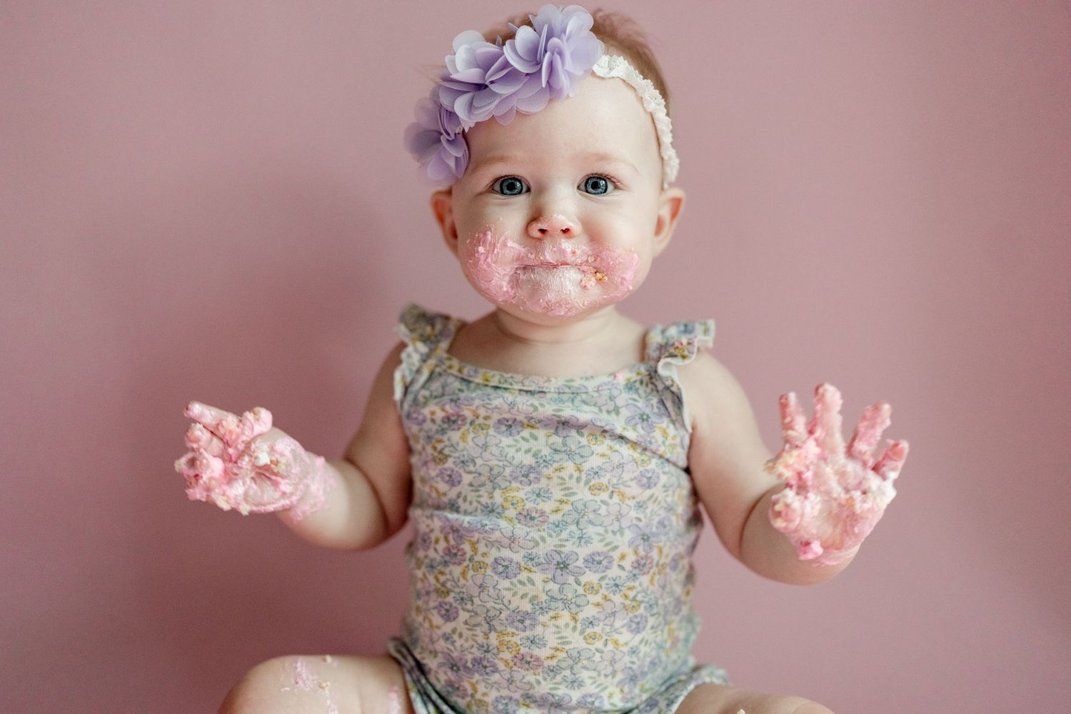 Baby covered in pink frosting shows messy hands while sitting next to birthday cake.