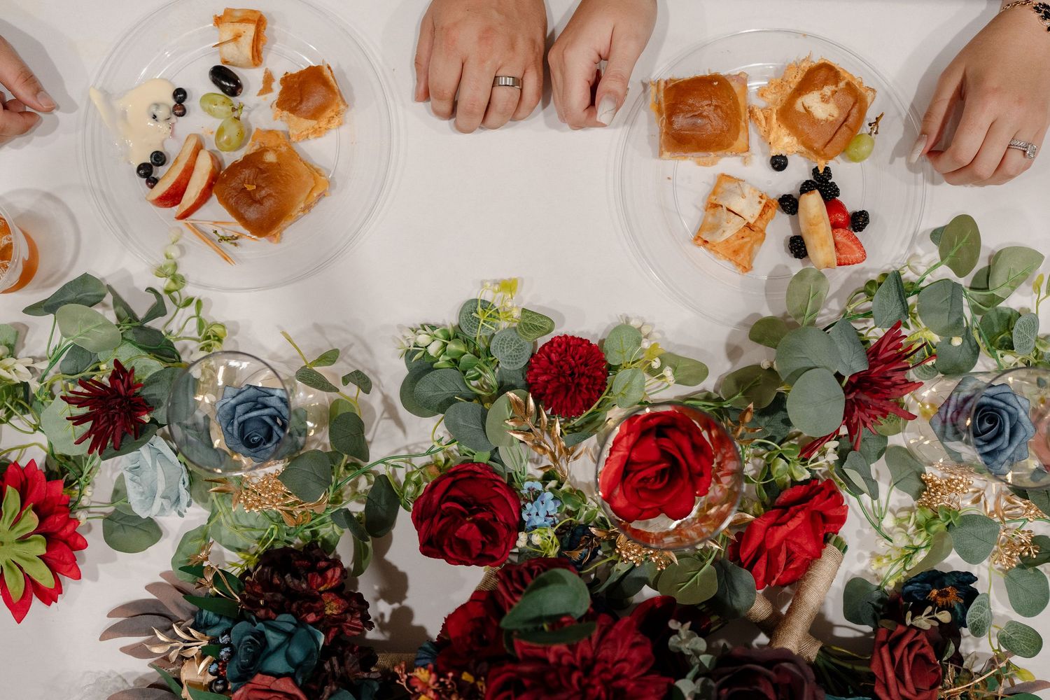 Hands touch colorful wedding flowers with red roses and blue accents arranged on a white surface.