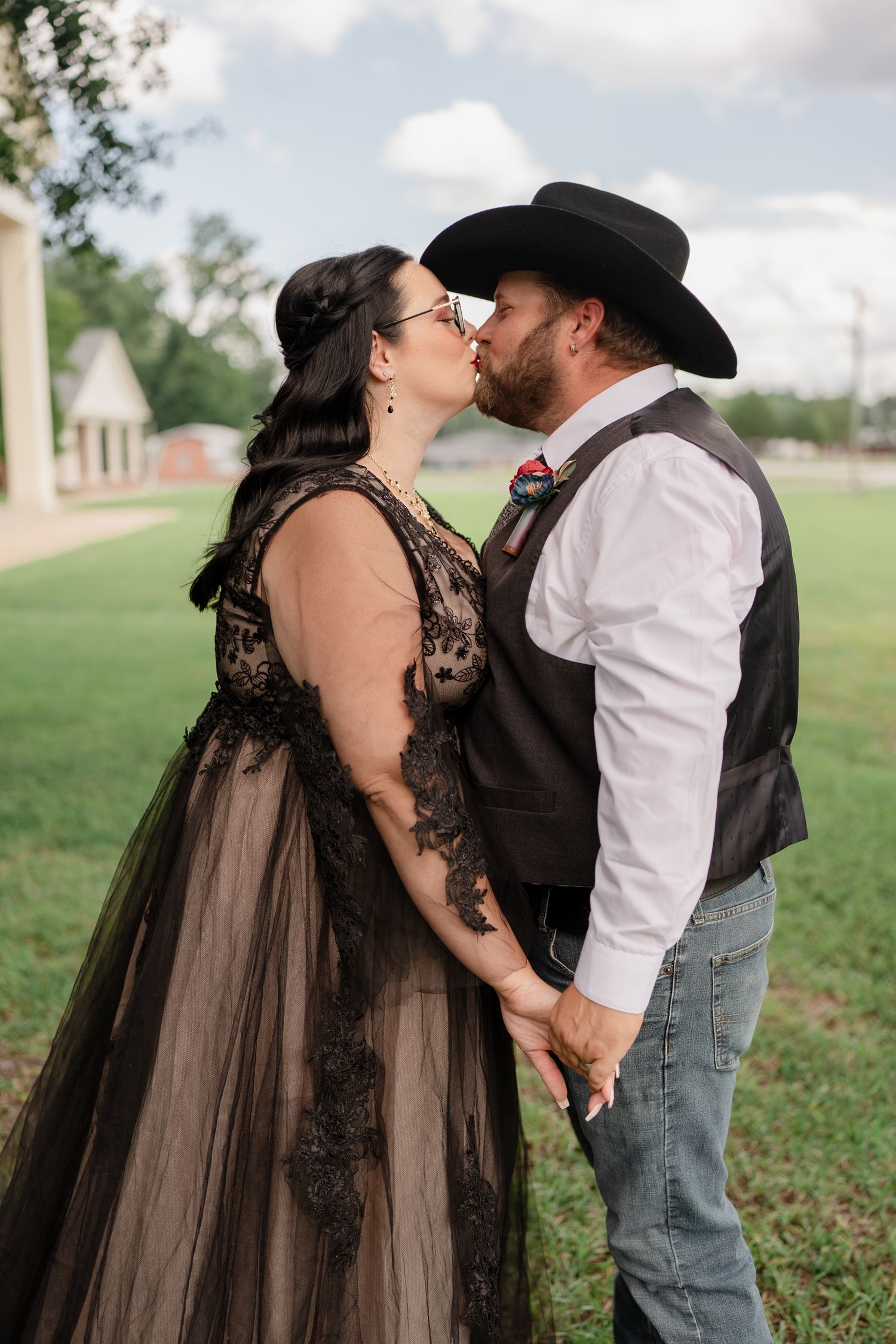 A couple in western attire share a romantic kiss in a grassy field on a cloudy day.