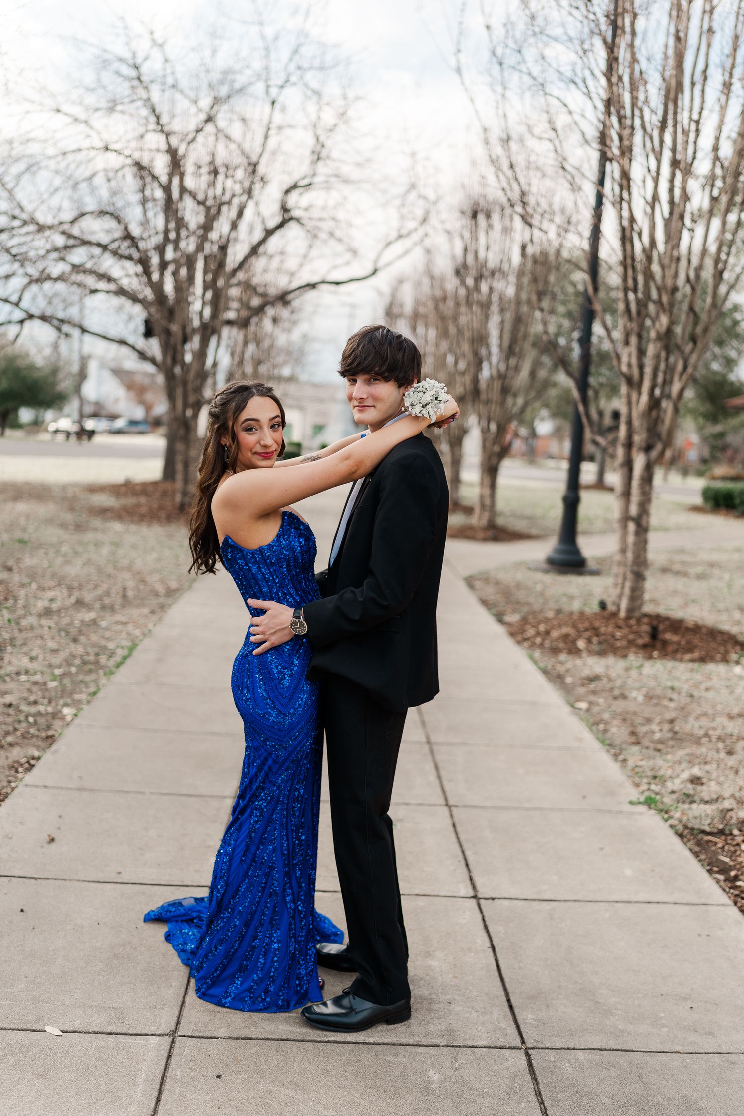 Couple in formal wear embraces on sidewalk surrounded by winter trees and lampposts.