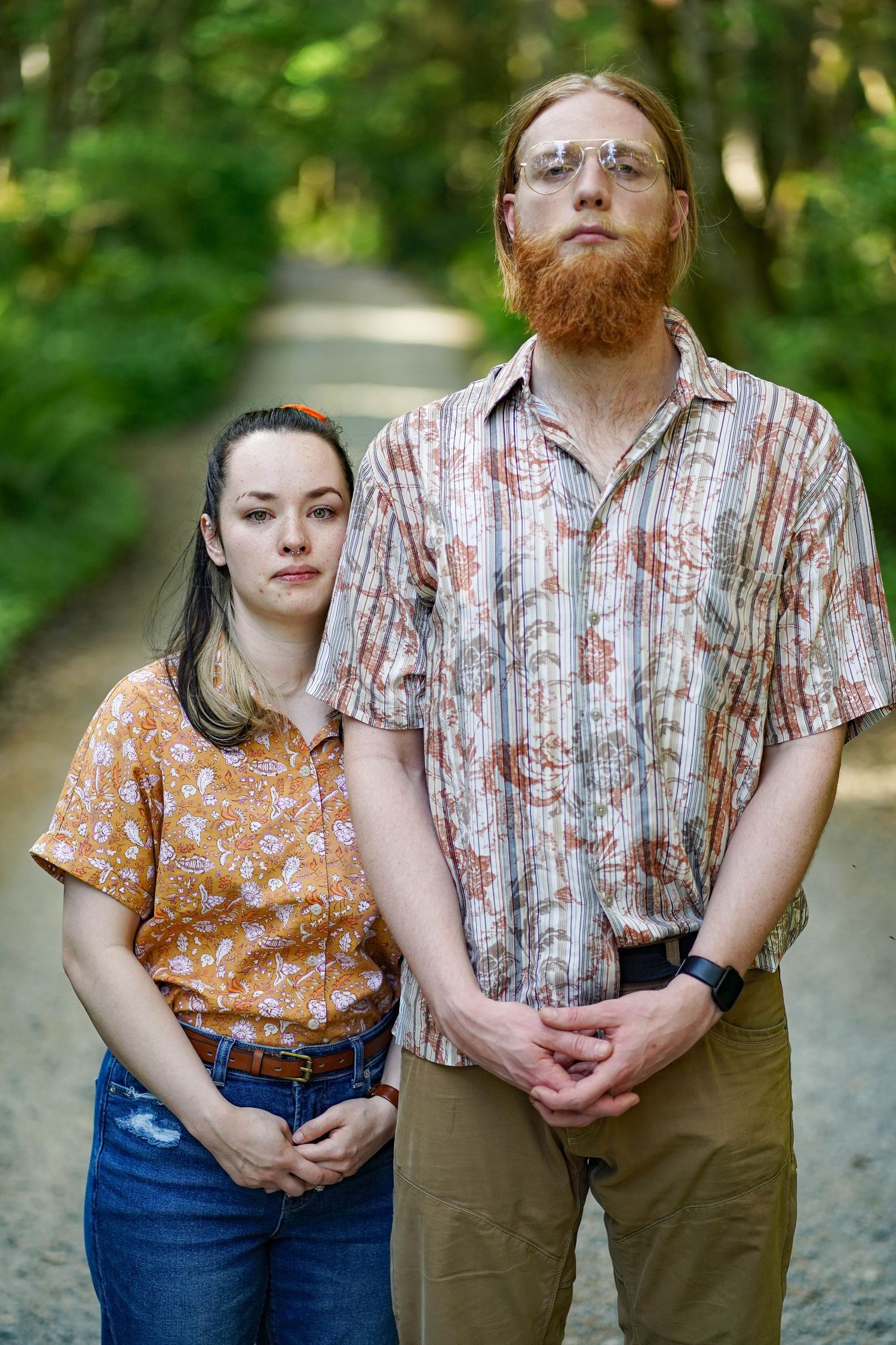 A height-contrasting portrait of two people in floral and striped shirts on a forest path.