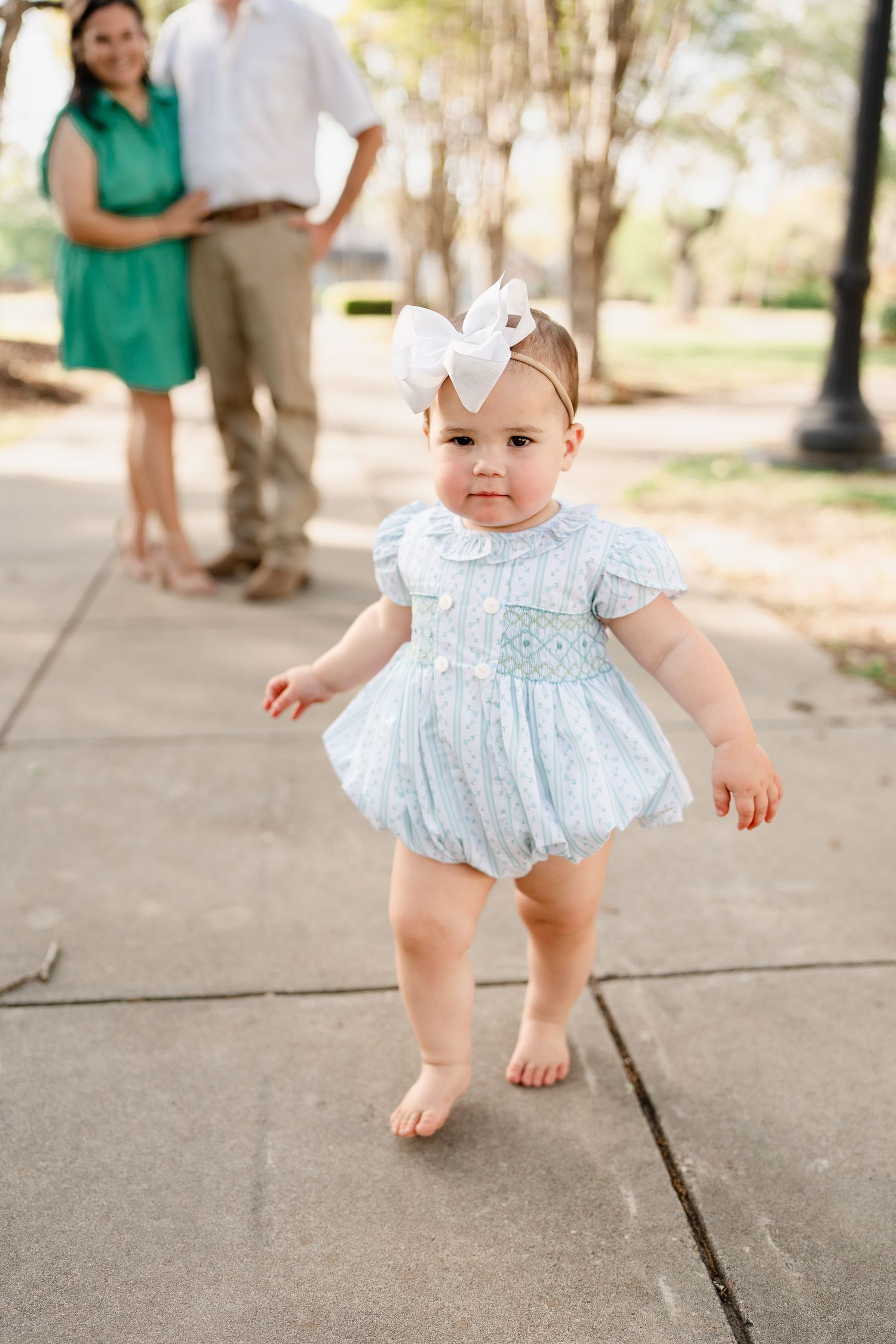 Baby in blue dress and white bow takes steps on concrete path with adults in background.